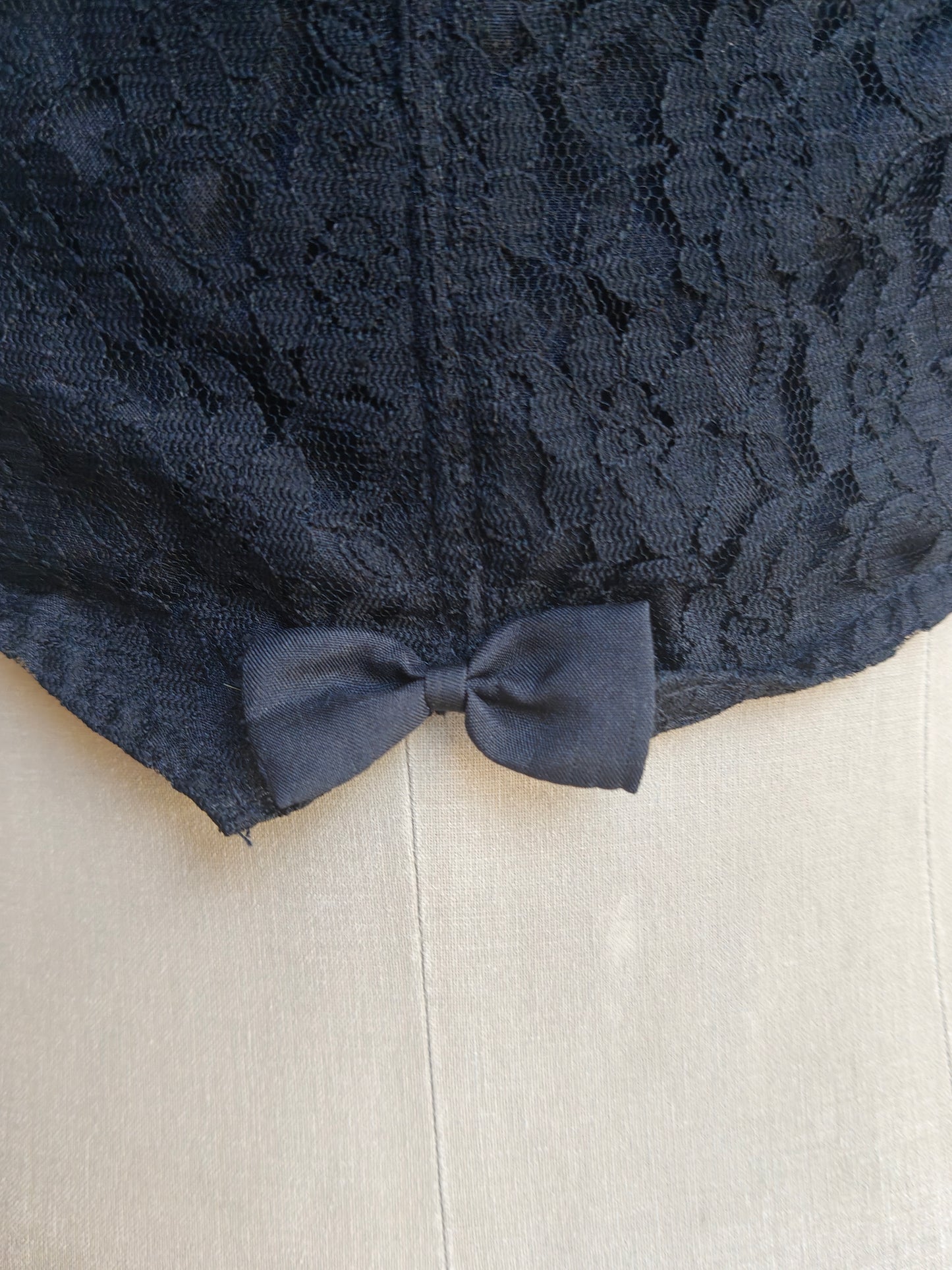 Close-up of the black lace back of top with a matching fabric bow at the hem, displayed on a light beige mannequin or dress form.