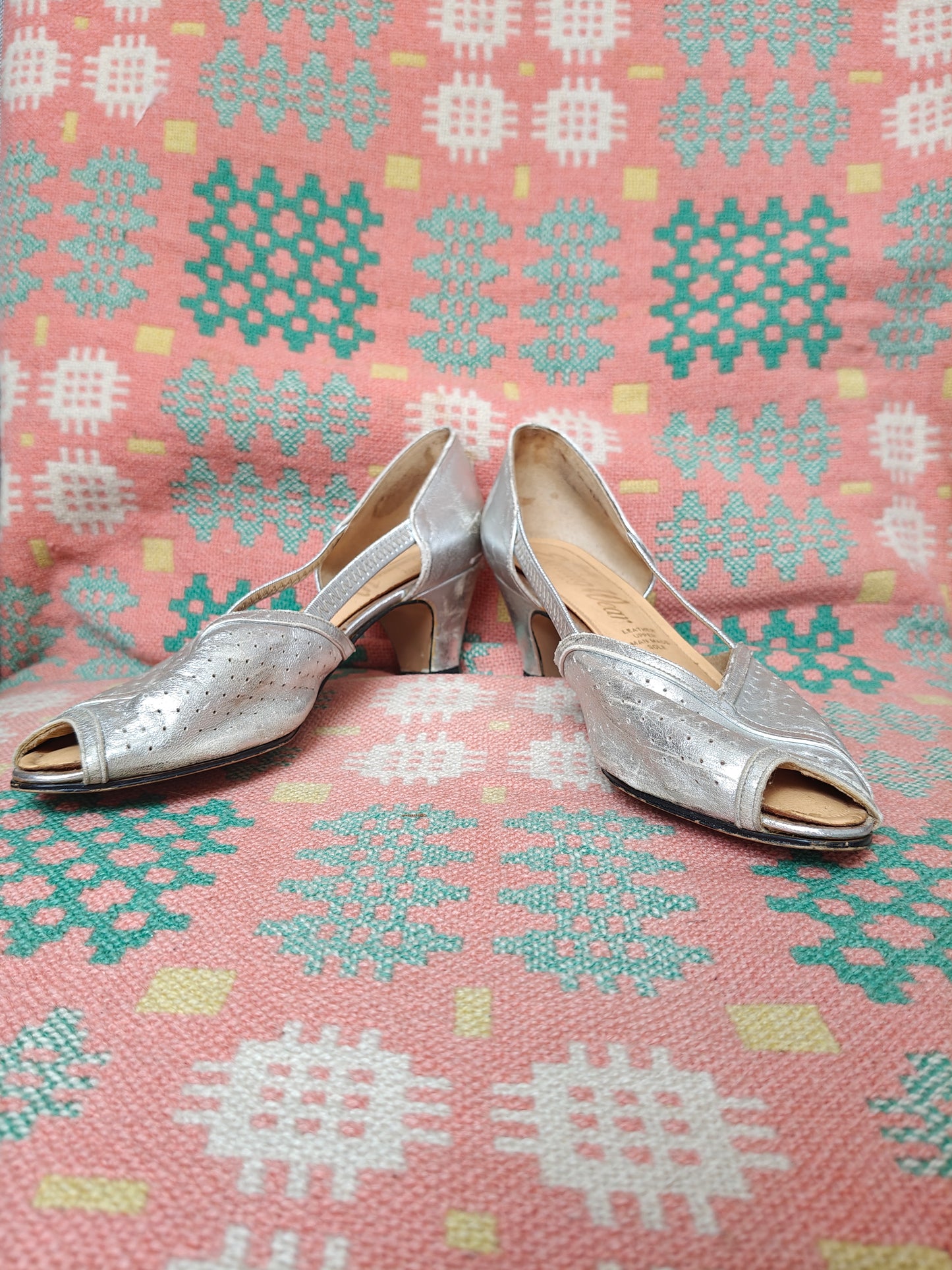 A pair of women's metallic silver peep-toe heels with low block heels, displayed on a pink and teal patterned woven blanket.