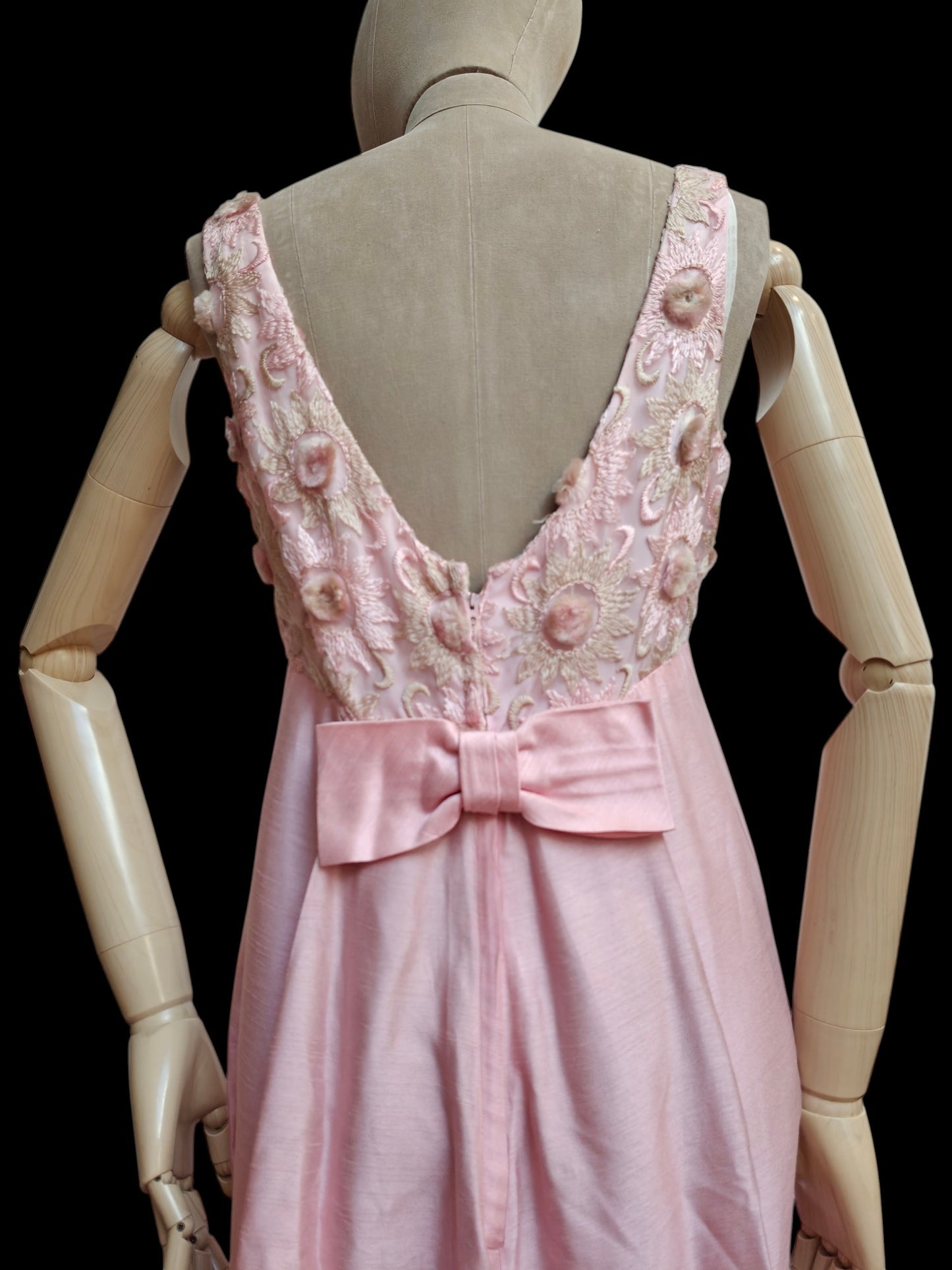 A light pink sleeveless dress displayed on a beige wooden mannequin, showing the back view with a deep V-neckline, floral applique, and a large satin bow at the waist against a black background.