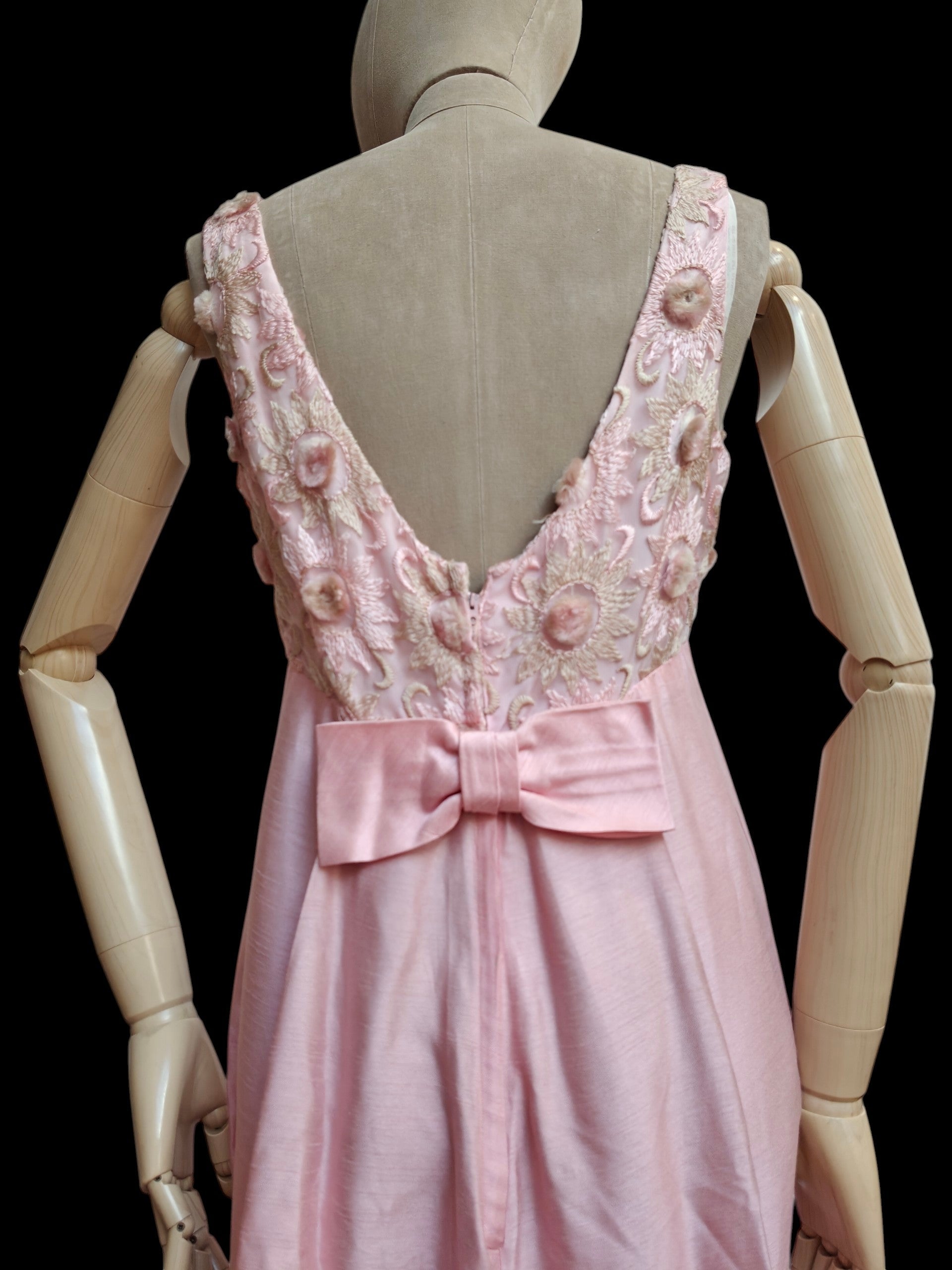 A light pink sleeveless dress displayed on a beige wooden mannequin, showing the back view with a deep V-neckline, floral applique, and a large satin bow at the waist against a black background.