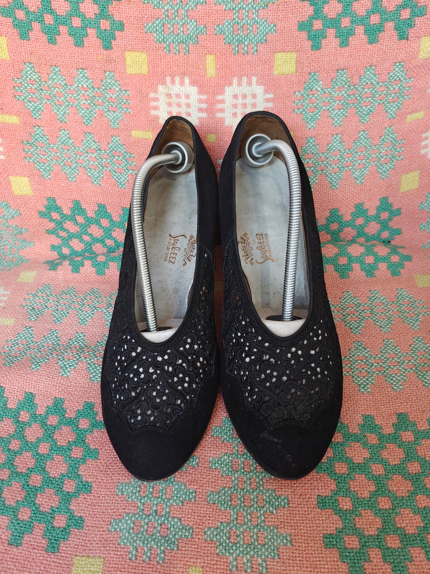 A pair of black slip-on shoes with decorative perforated detailing on the sides, shown on a patterned pink and green textile background.