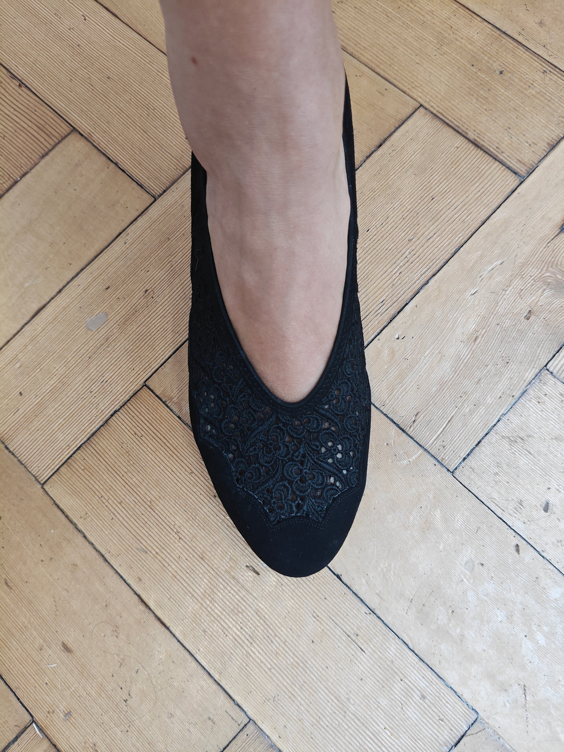 A person's foot wearing a black slip-on shoe with a lace-embroidered toe panel, standing on a light-colored wooden parquet floor.