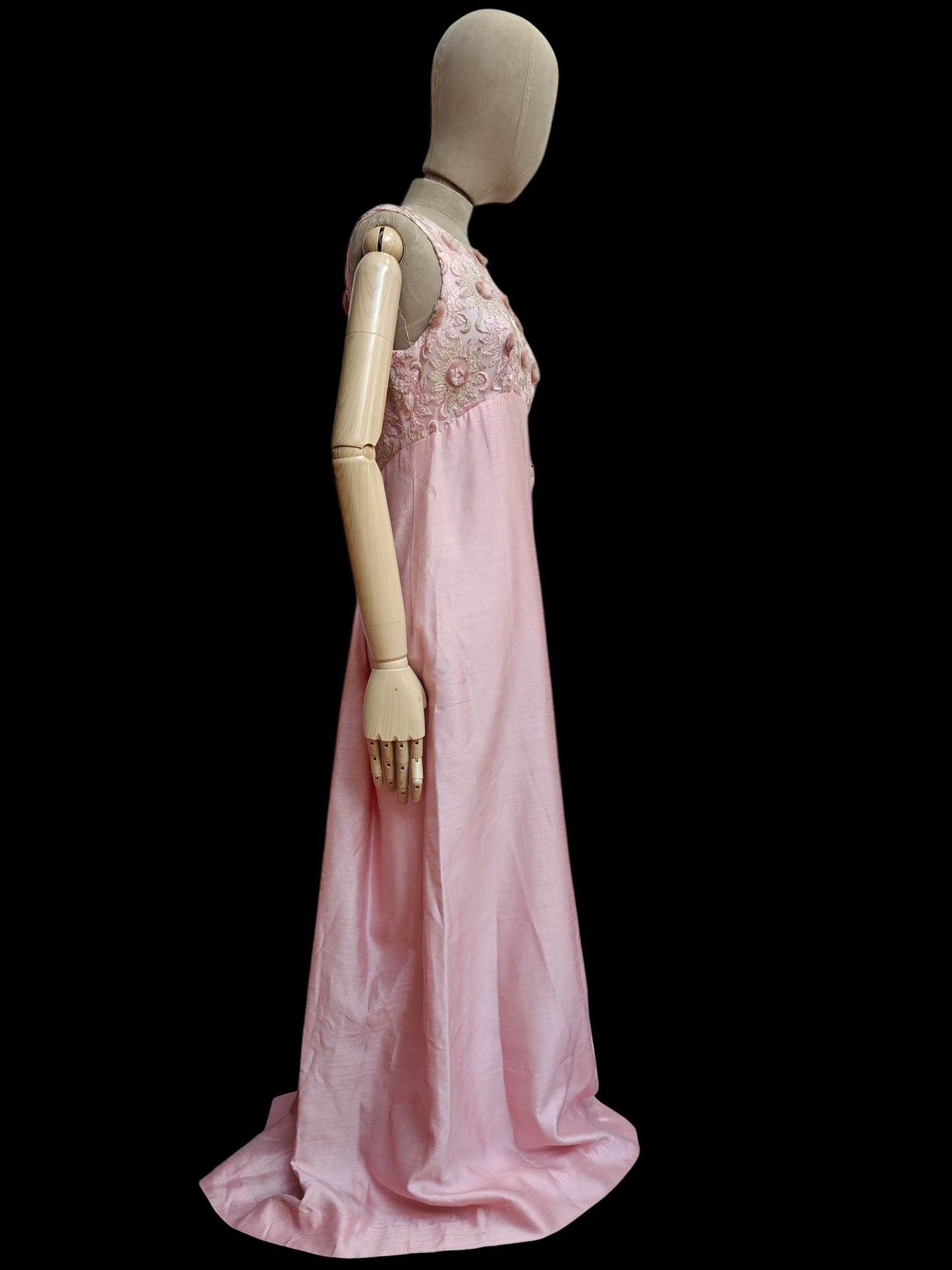 A long light pink vintage gown displayed on a beige mannequin against a solid black background. The sleeveless dress features a heavily embroidered floral bodice.