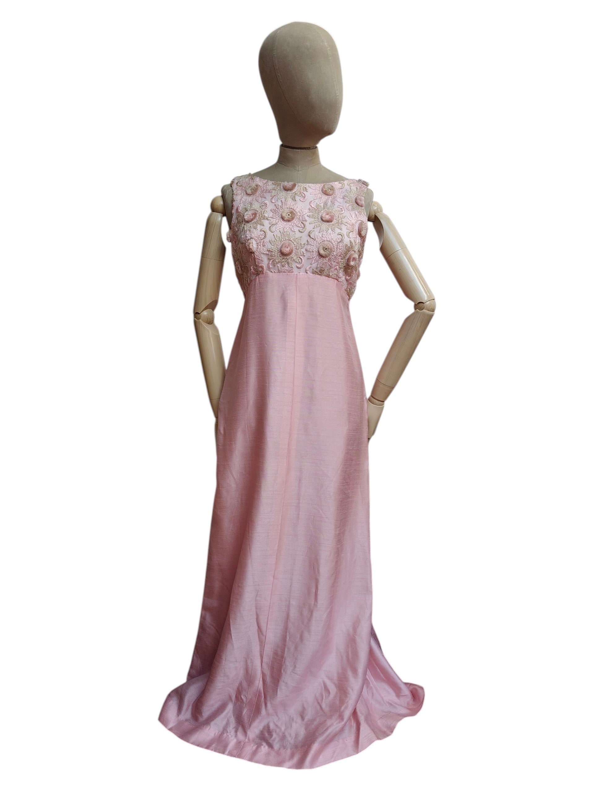 A pink sleeveless vintage maxi dress displayed on a wooden mannequin against a plain white background. The dress features a textured pink skirt and a heavily embellished bodice with pink and white floral embroidery and beadwork.