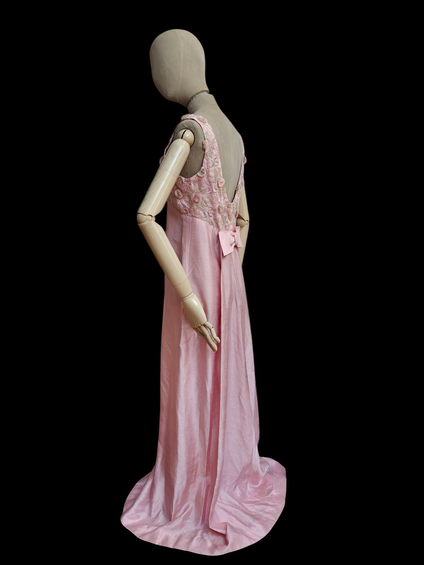 A pink floor-length gown displayed on a beige mannequin against a solid black background. The dress features a fitted bodice with floral embroidery and a bow at the back, with a flowing silk-like skirt.