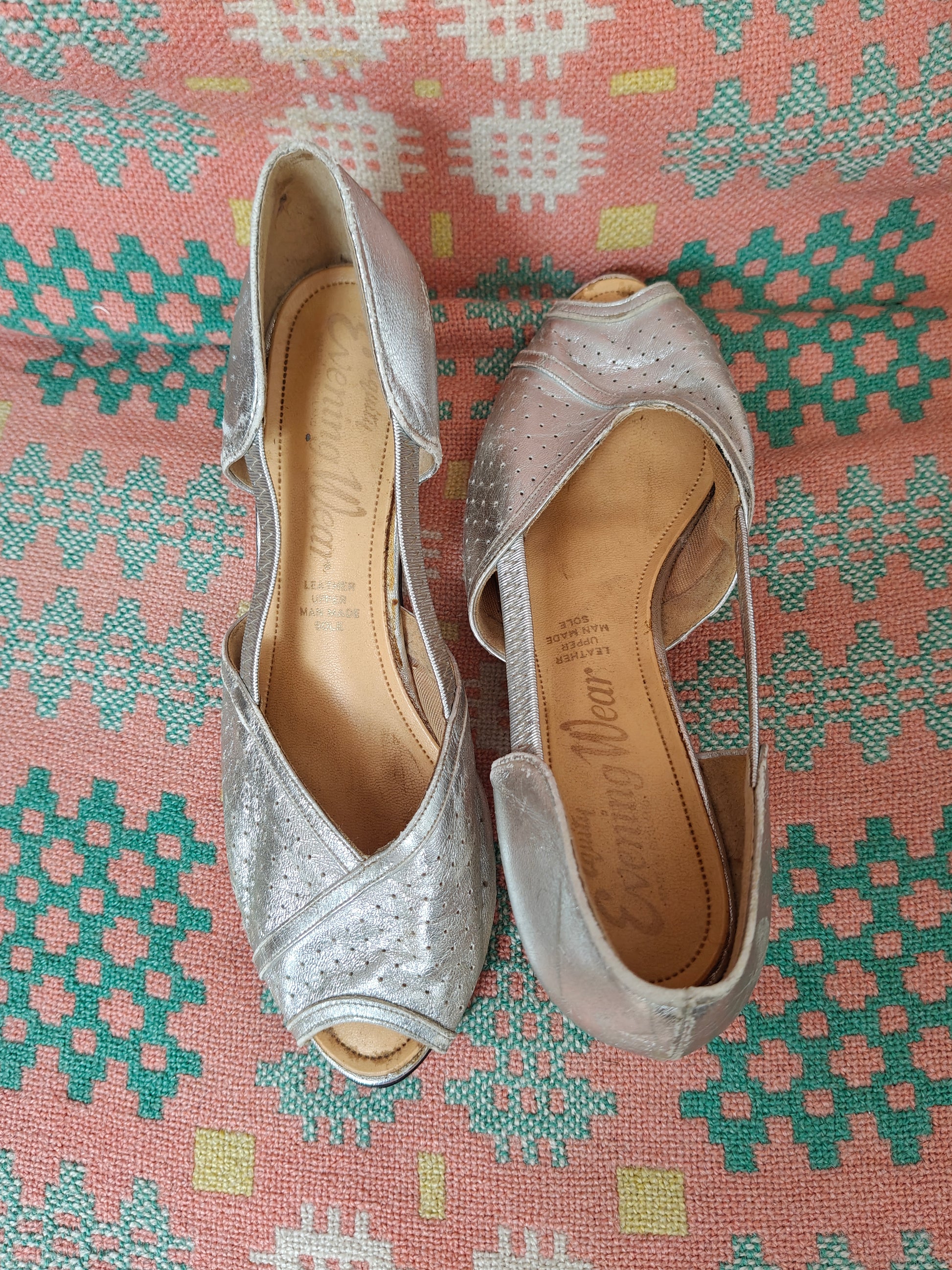 A pair of women's peep-toe  heeled sandals in metallic silver with perforated detailing, displayed on a pink and green patterned textile background. The brand 'Evening Wear' is visible on the tan insoles.