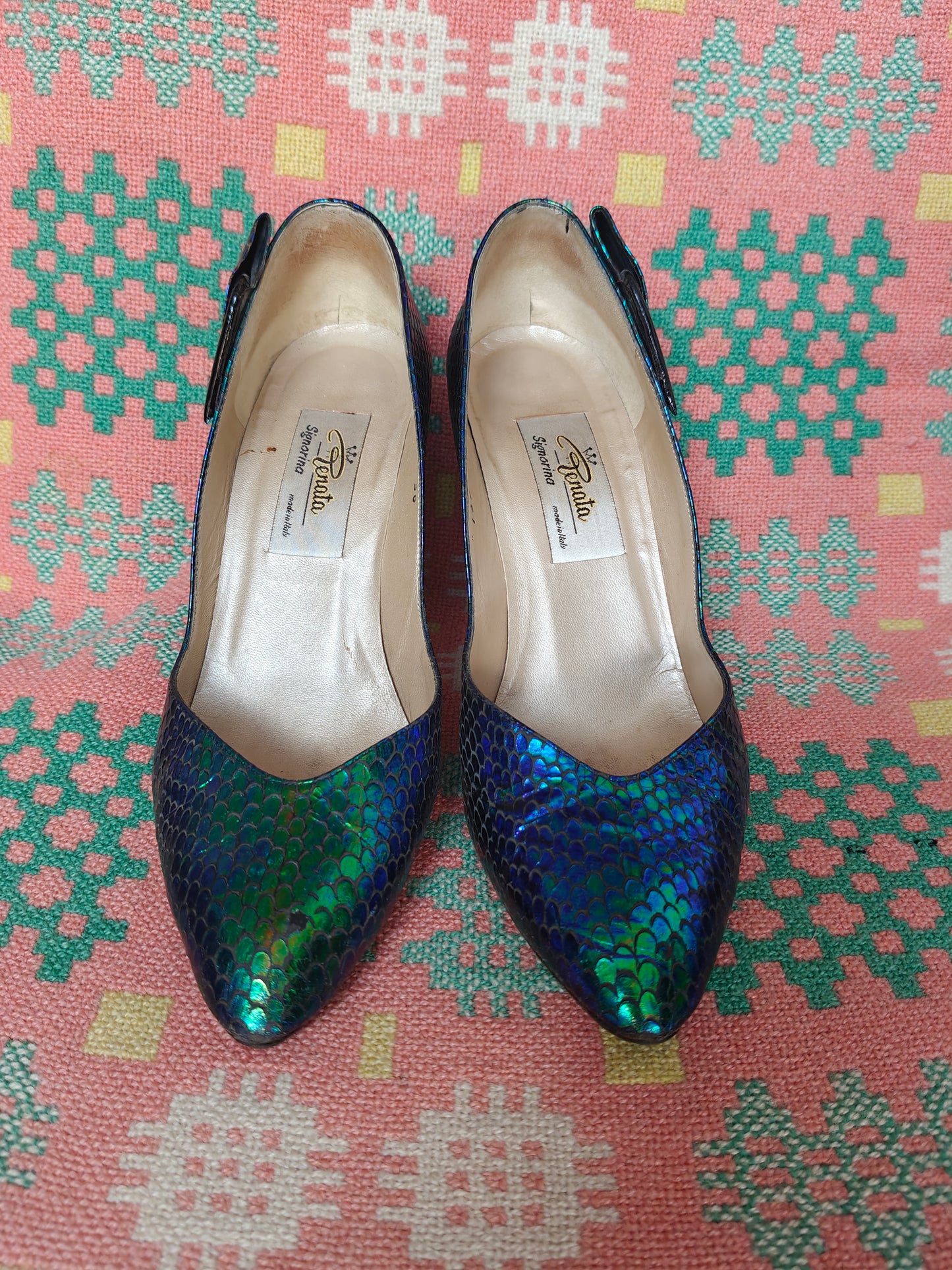 A pair of pointed-toe heels with a metallic, iridescent green and blue scale pattern, resting on a pink patterned textile background. The brand name Renata is visible on the insoles.
