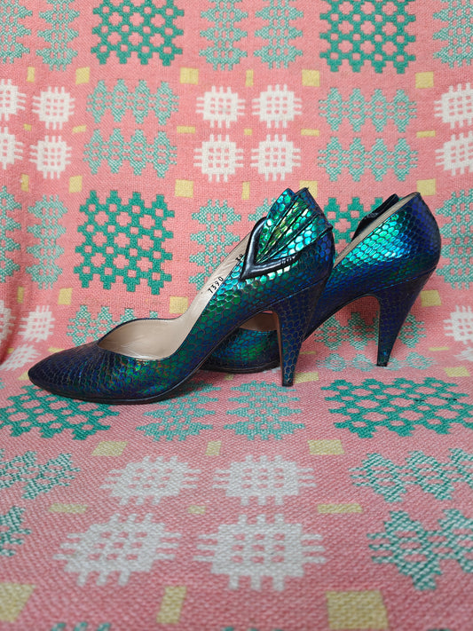 A pair of iridescent blue and green high-heeled pumps with a snakeskin texture, displayed on a pink and teal patterned textile background.