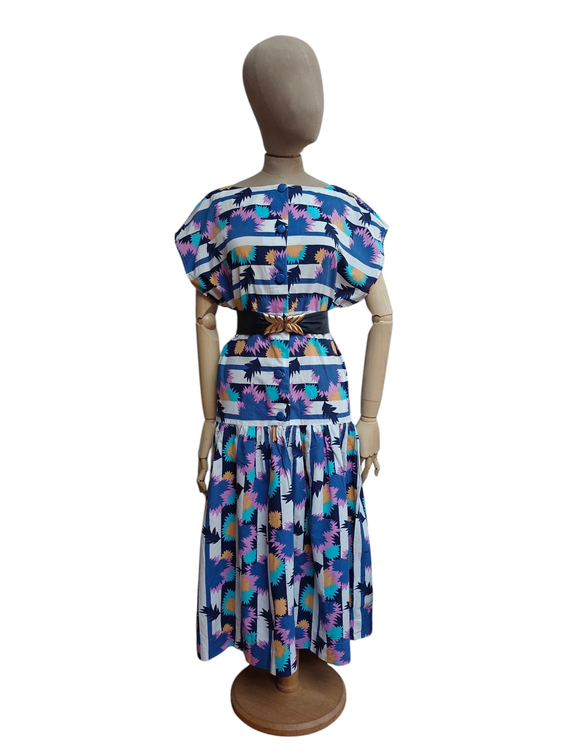 A vintage blue patterned dress displayed on a wooden mannequin from the back, featuring a blue and white striped bodice with colorful floral motifs and a matching flared skirt.