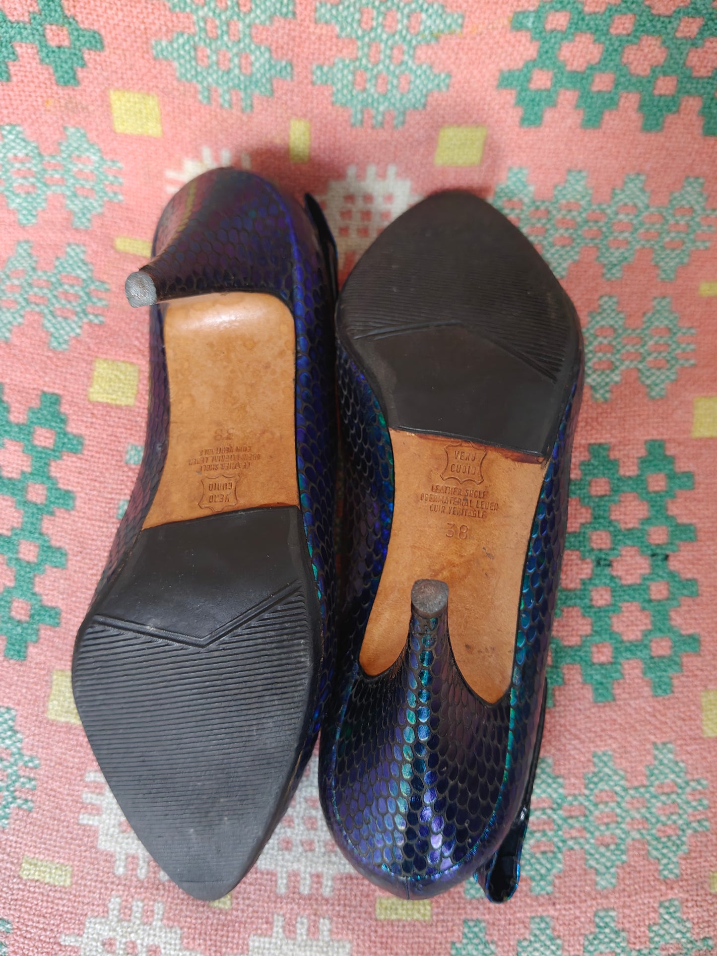 A pair of women's pointed-toe flats with a dark blue and black snakeskin pattern, shown from both the top and bottom sides on a pink and green patterned textile background.