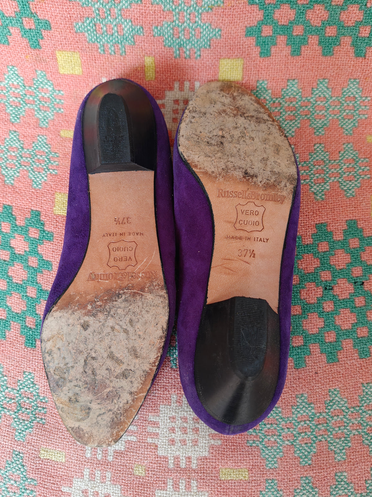 A pair of purple suede flat shoes shown from the bottom, revealing heavily worn and scuffed soles, placed on a pink and green patterned textile background.