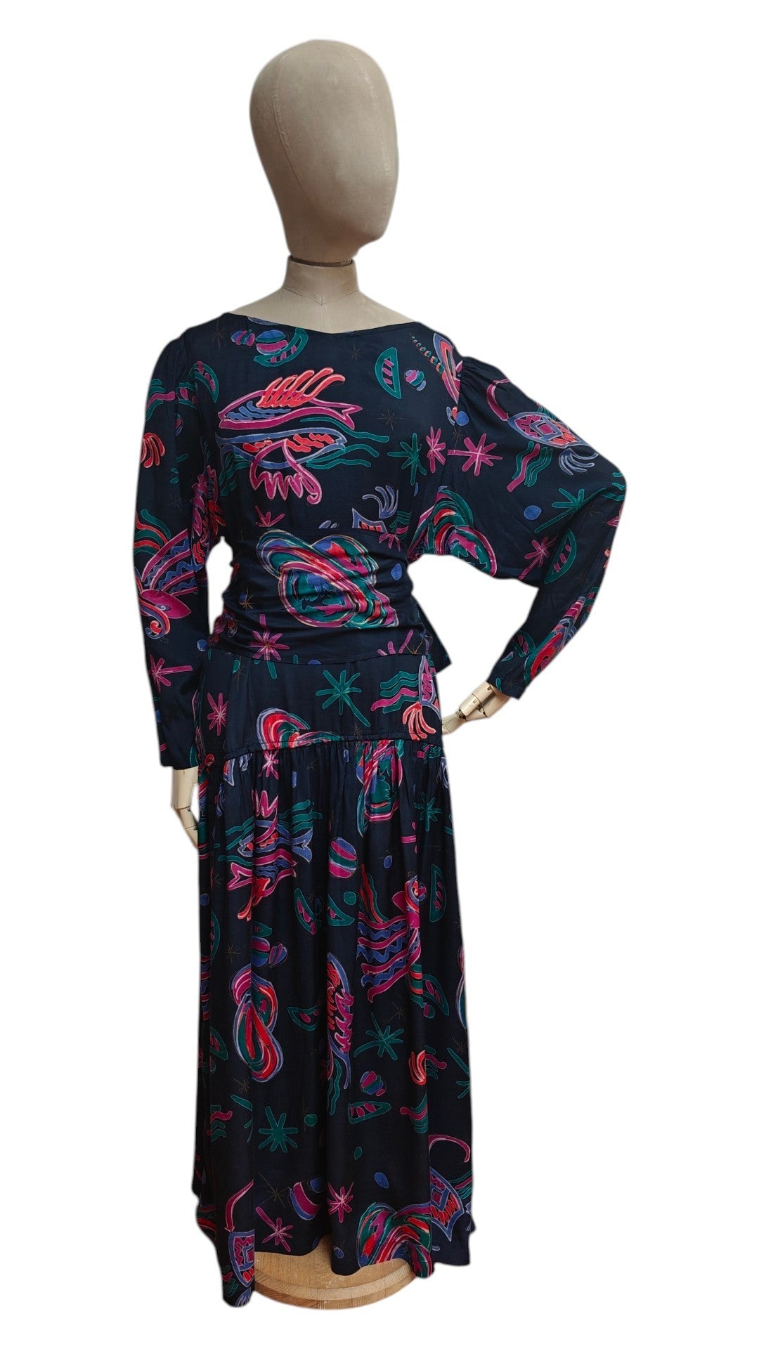 A long-sleeve maxi dress displayed on a mannequin from the back, featuring a black base with a vibrant pink, teal, and red abstract paisley pattern.