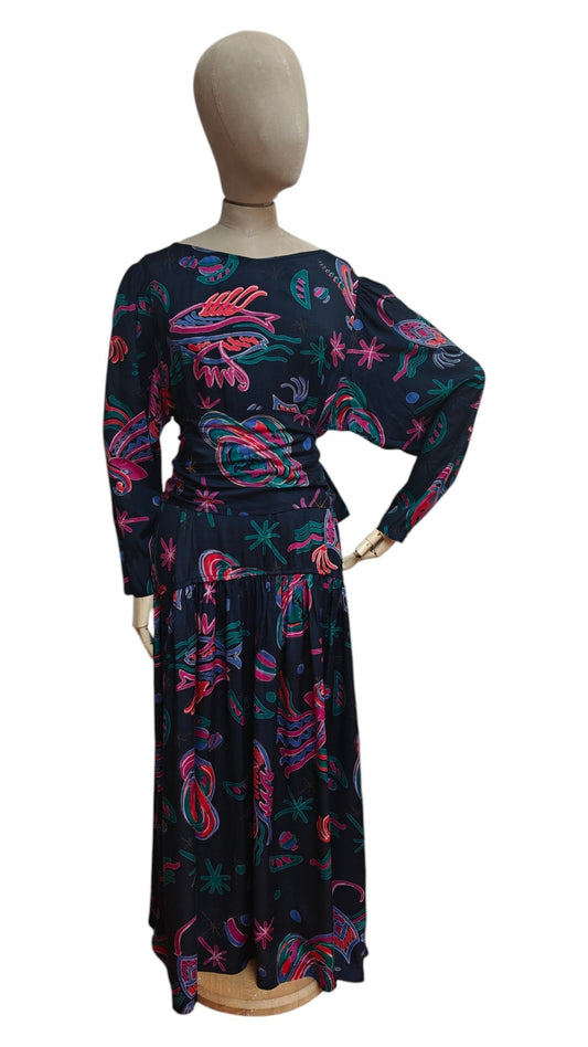 A long-sleeve maxi dress displayed on a mannequin from the back, featuring a black base with a vibrant pink, teal, and red abstract paisley pattern.