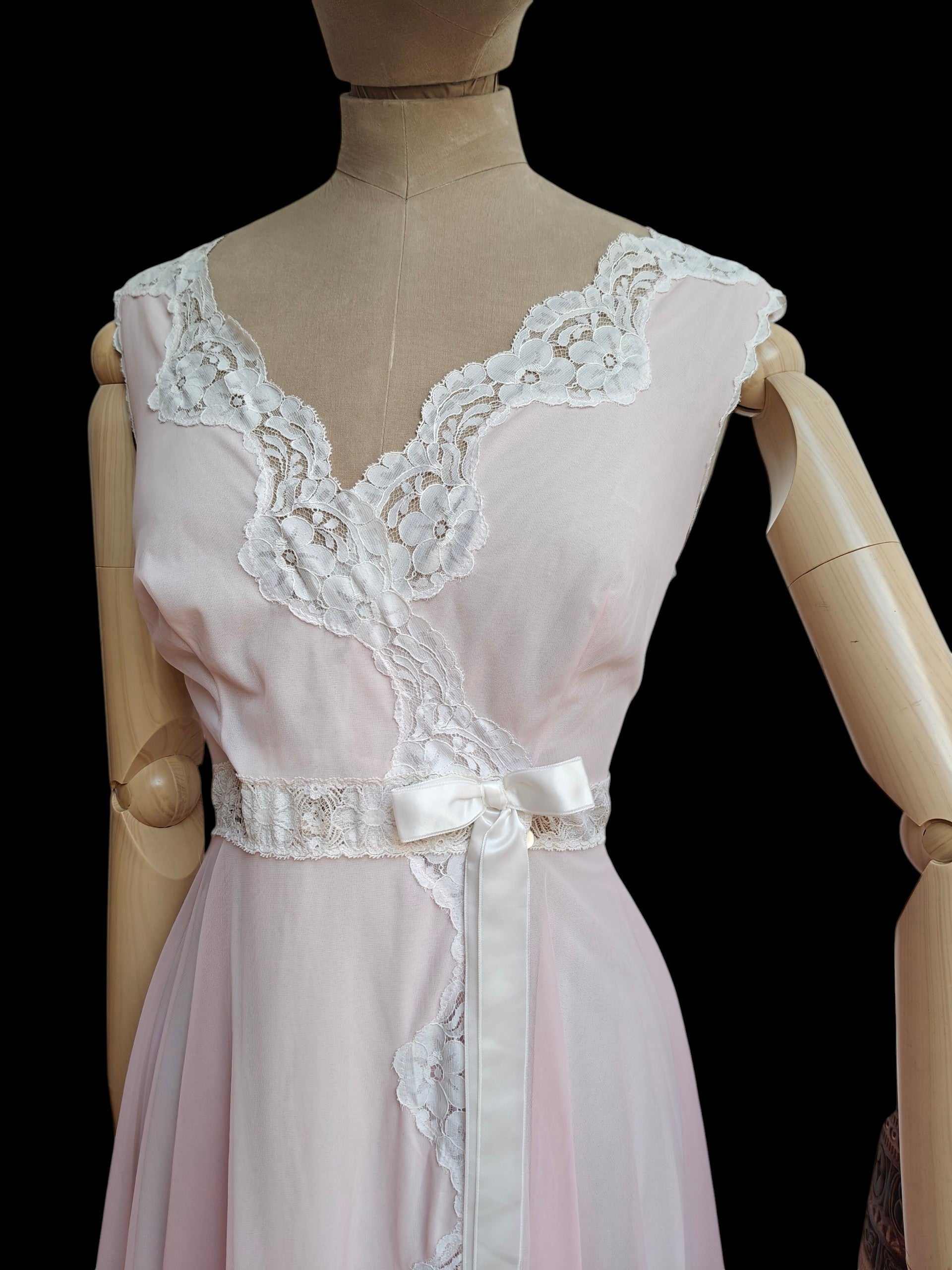 A light pink vintage nightgown with white lace trim and a matching white satin waist tie, displayed on a wooden mannequin against a black background.