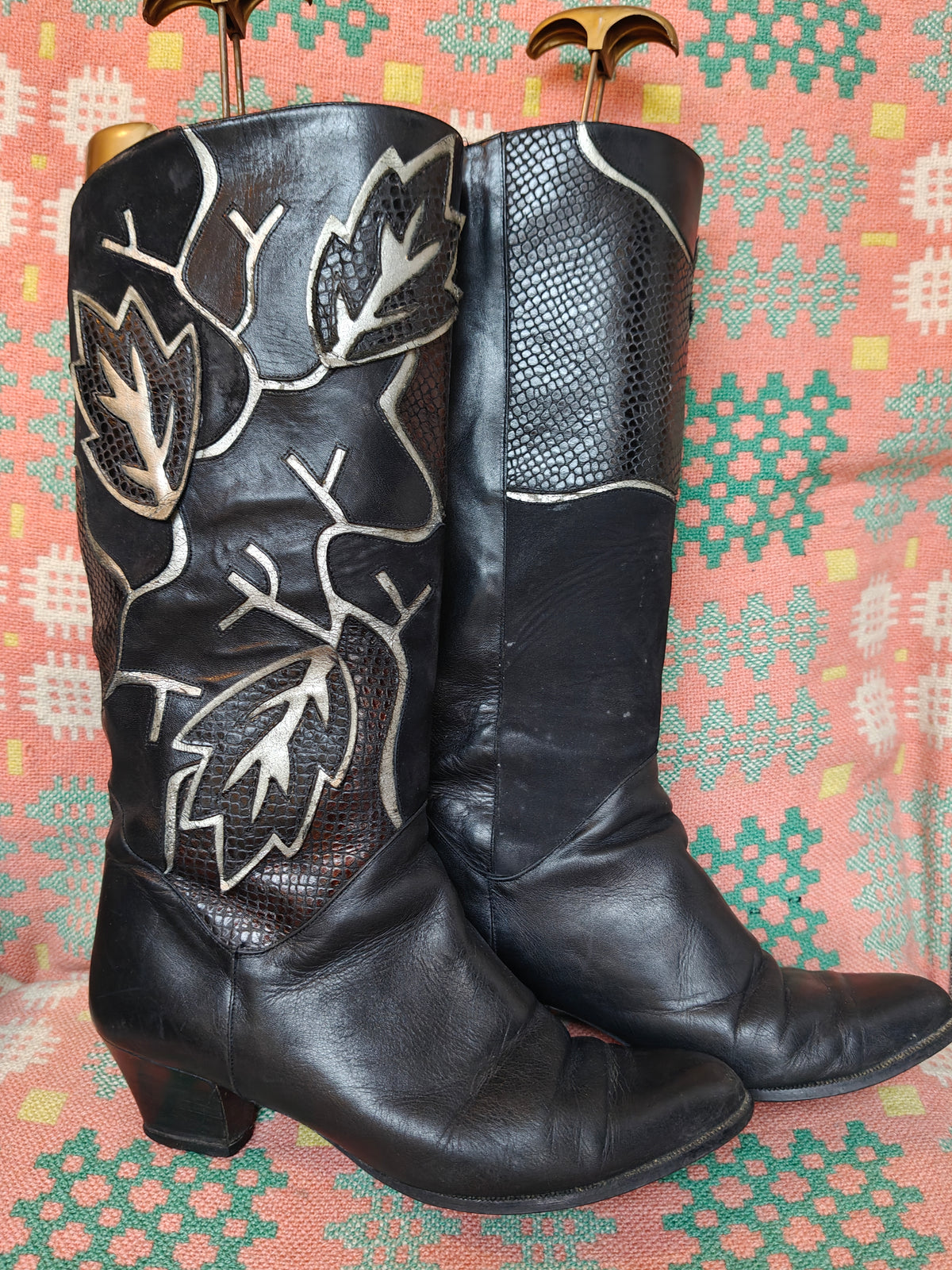 A pair of black leather knee-high cowboy boots with textured snakeskin and smooth leather panels, featuring white embroidered leaf designs, displayed on wooden hangers against a patterned pink and green fabric background.