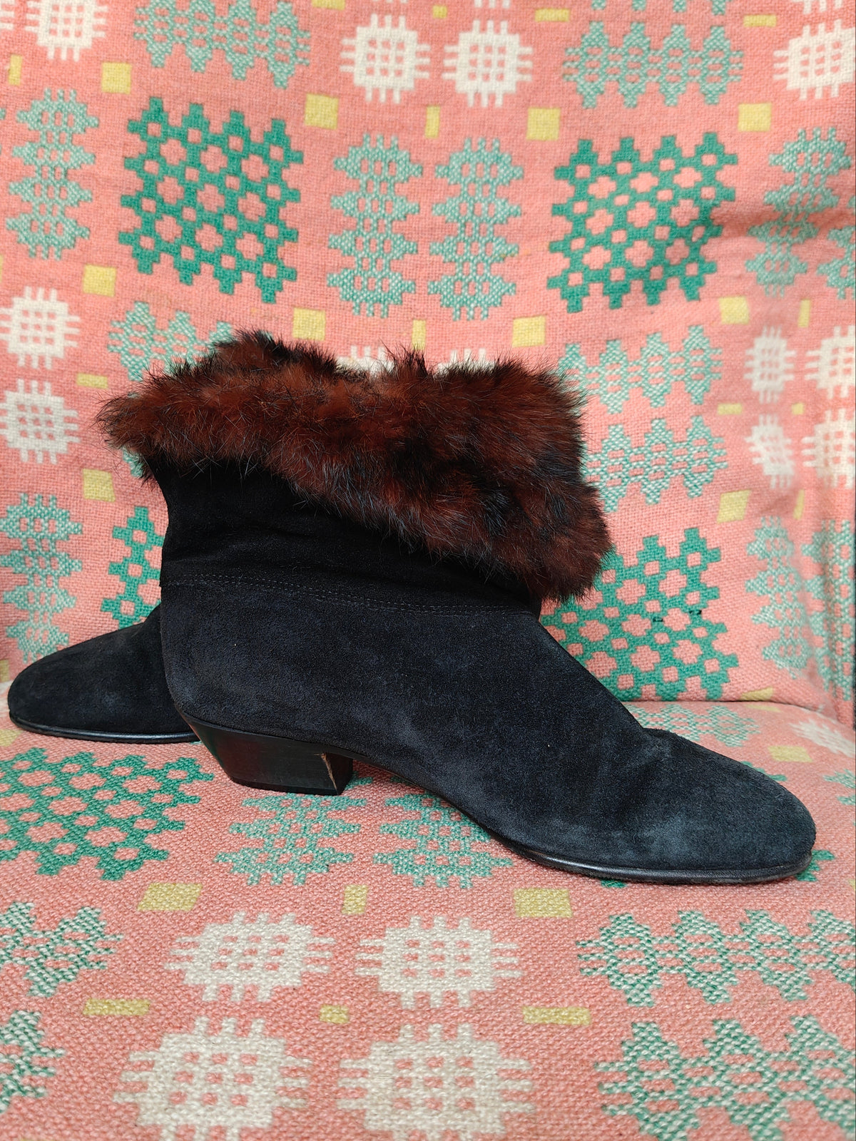 A pair of black suede ankle boots with dark brown faux fur trim, resting on a pink and green patterned woven blanket.
