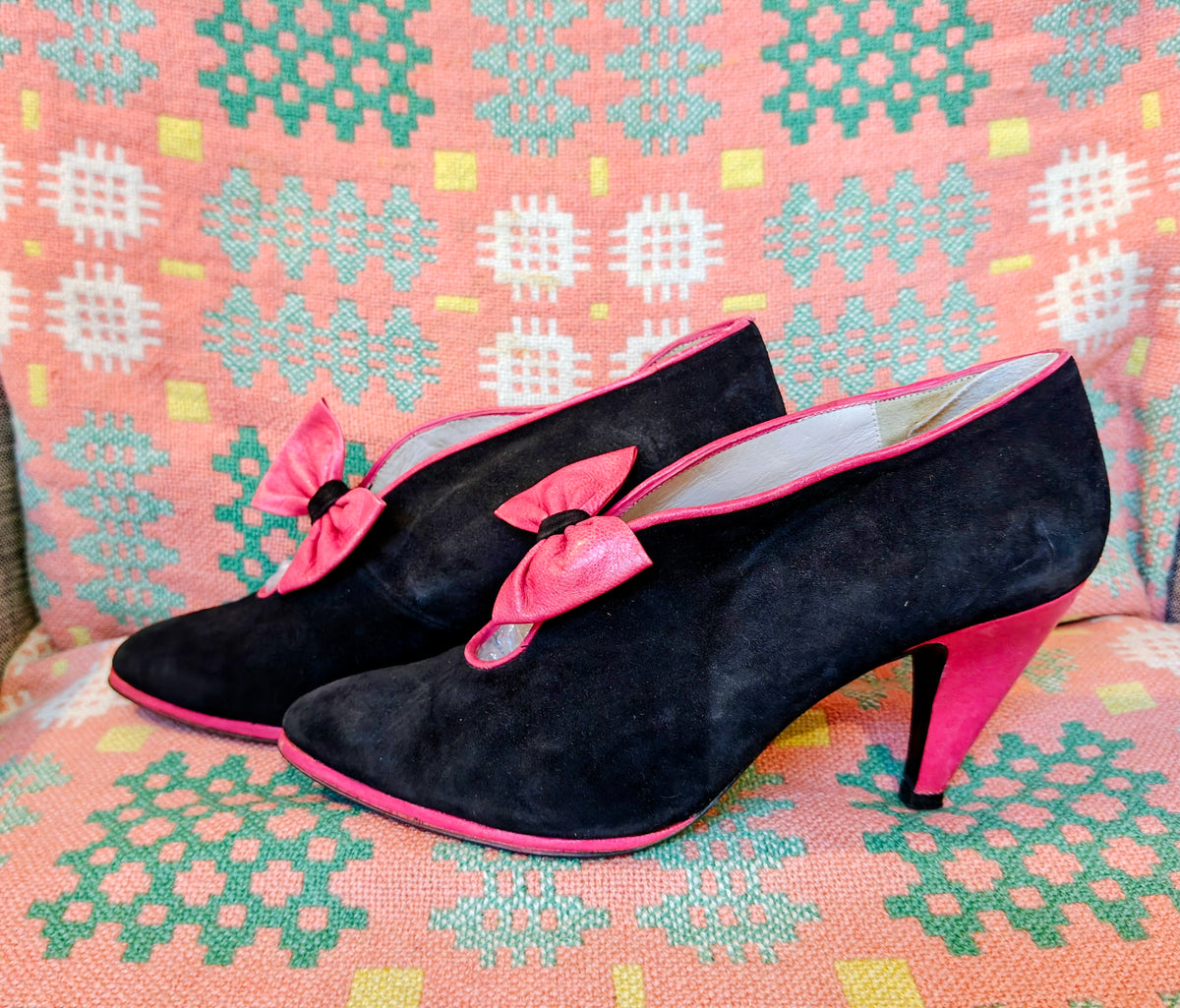 A pair of black suede high-heeled shoes with bright pink bows and pink trim, displayed on a patterned pink, green, and white textile background.