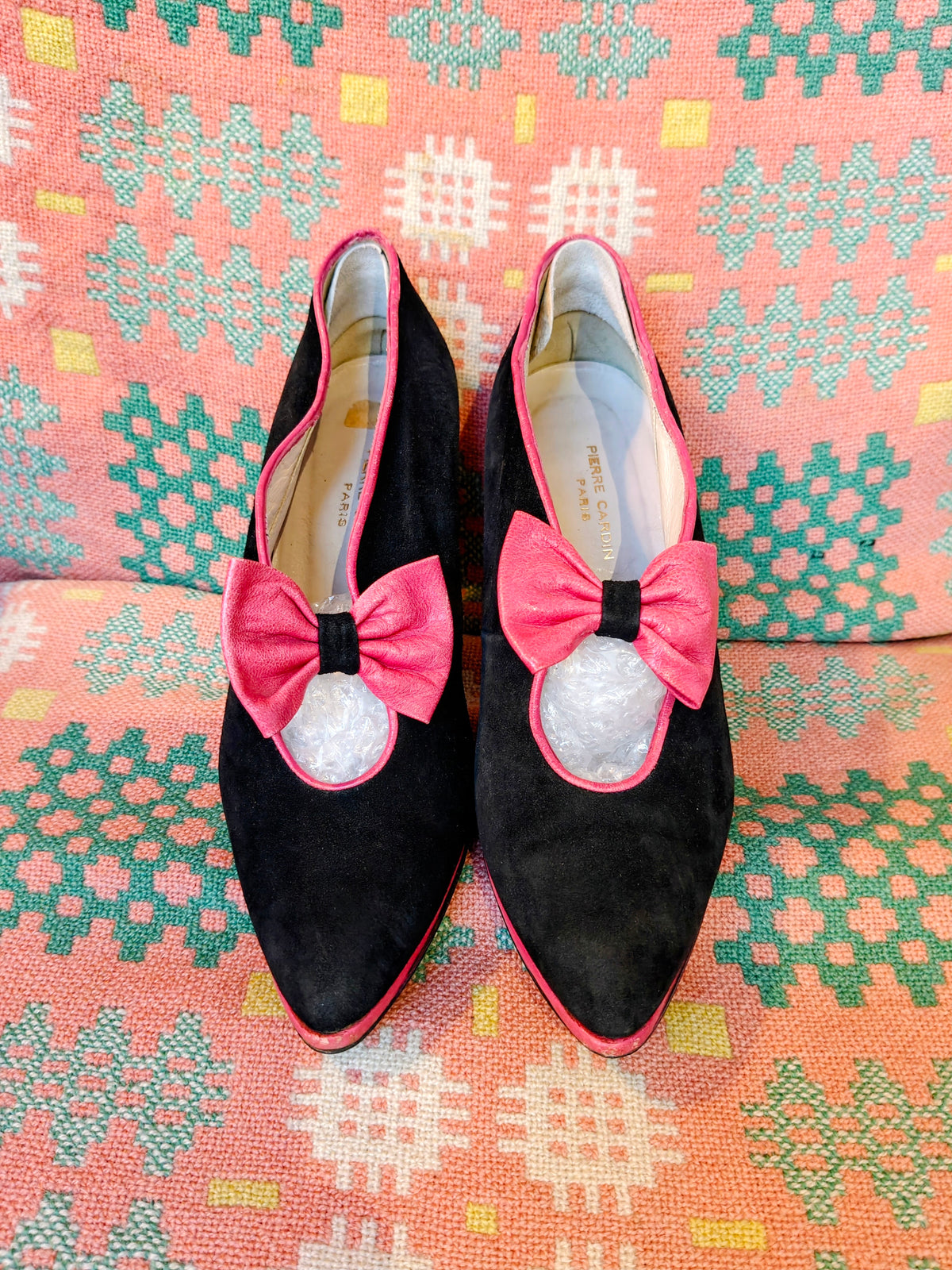 A pair of black suede pointed-toe flats with bright pink bow detail displayed on a  patterned textile background.
