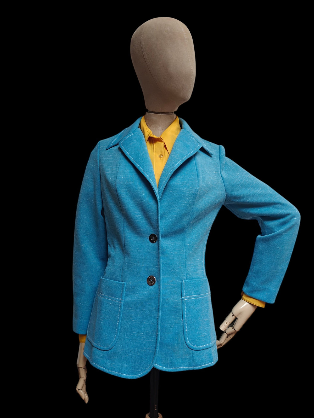 A bright blue 1970s blazer displayed on a beige mannequin torso, layered over a yellow collared shirt, set against a solid black background.