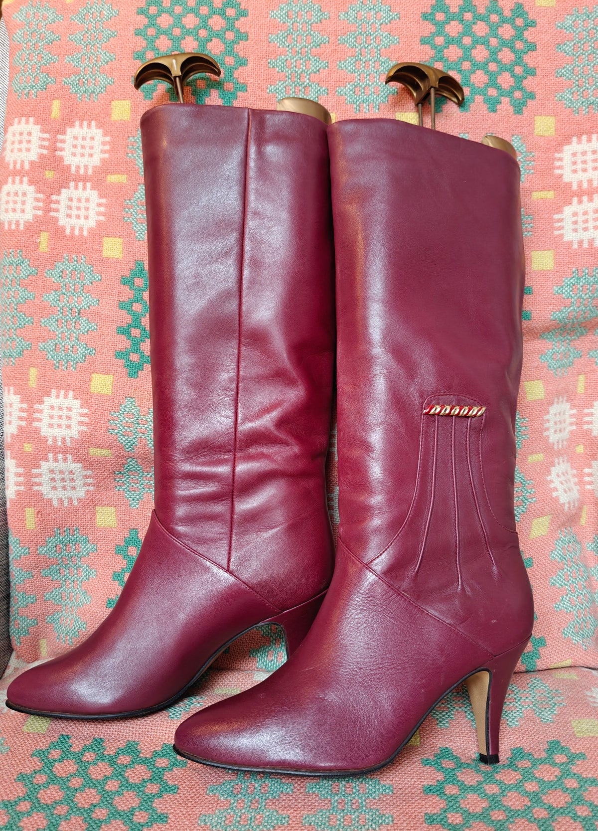 A pair of knee-high burgundy leather boots with pointed toes and heels displayed on patterned fabric.