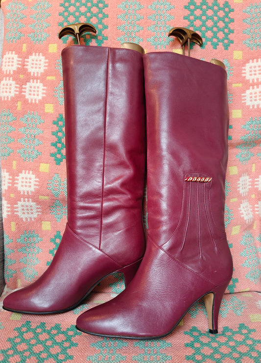 A pair of knee-high burgundy leather boots with pointed toes and heels displayed on patterned fabric.