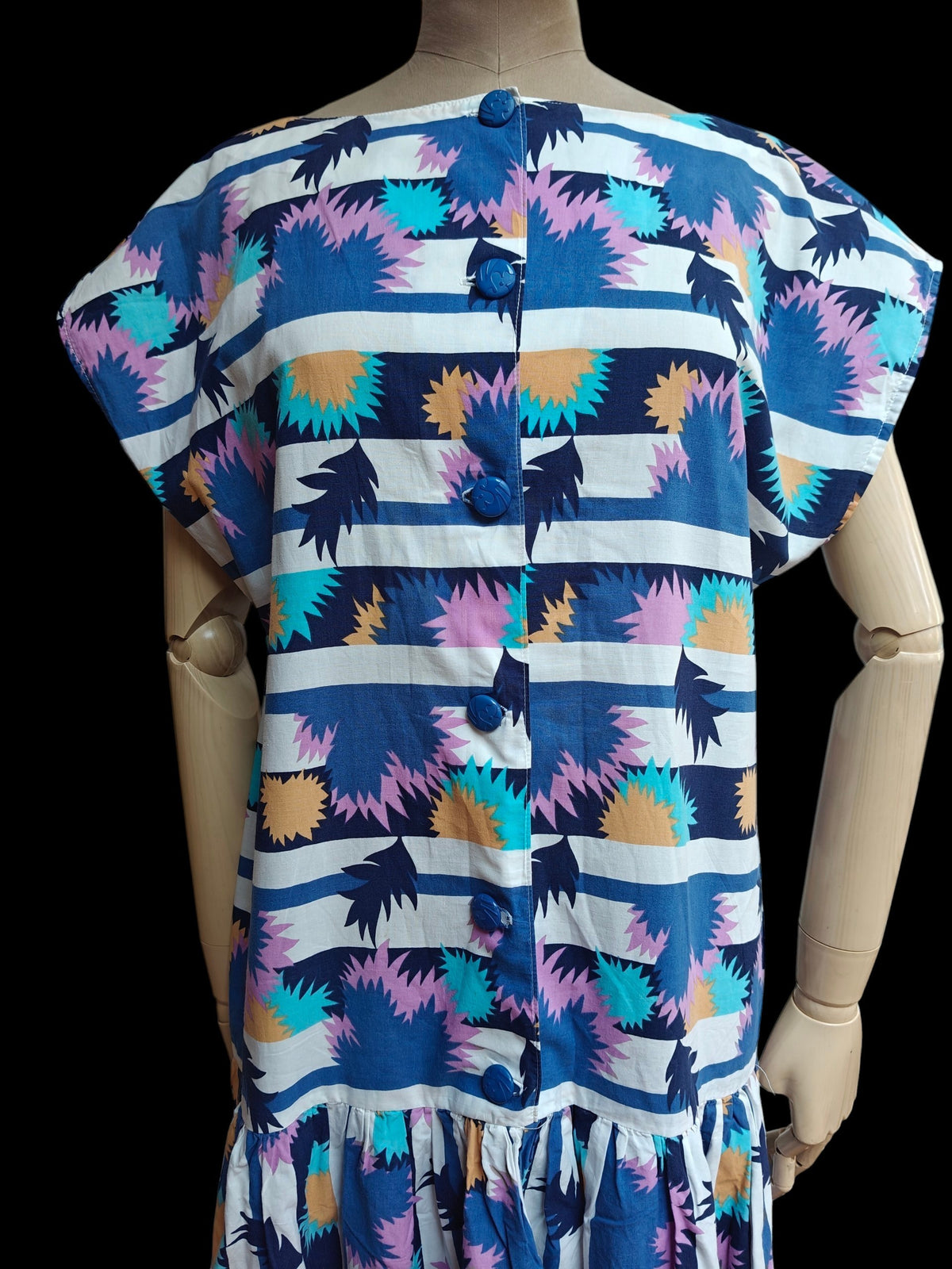 A short-sleeve button-front dress displayed on a wooden mannequin against a black background. The dress has a bold abstract pattern with horizontal navy, white, and blue stripes, accented with pink, teal, and mustard yellow shapes.