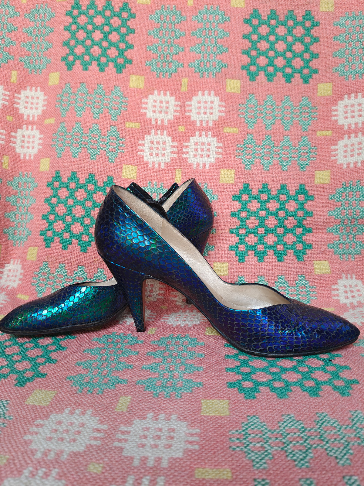 A pair of pointed-toe 80's high heels with a metallic blue and green iridescent snakeskin pattern, displayed on a pink patterned fabric background.
