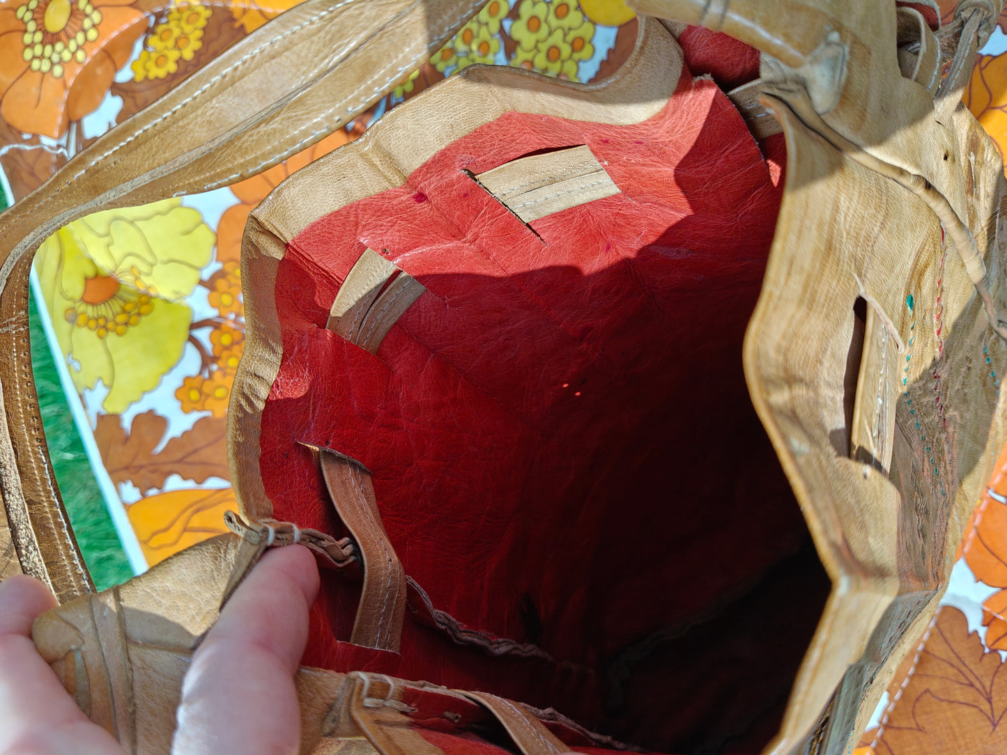 A close-up view of a vintage tan leather tote bag's interior, showing a red fabric lining and a hand holding the zipper tab on the left, set against a colorful floral background.