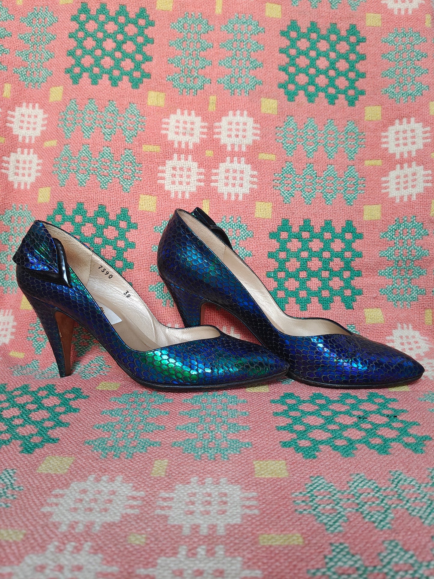 A pair of vintage pointed-toe high heels with a metallic iridescent blue and green scale pattern, displayed on a pink and teal patterned textile background.
