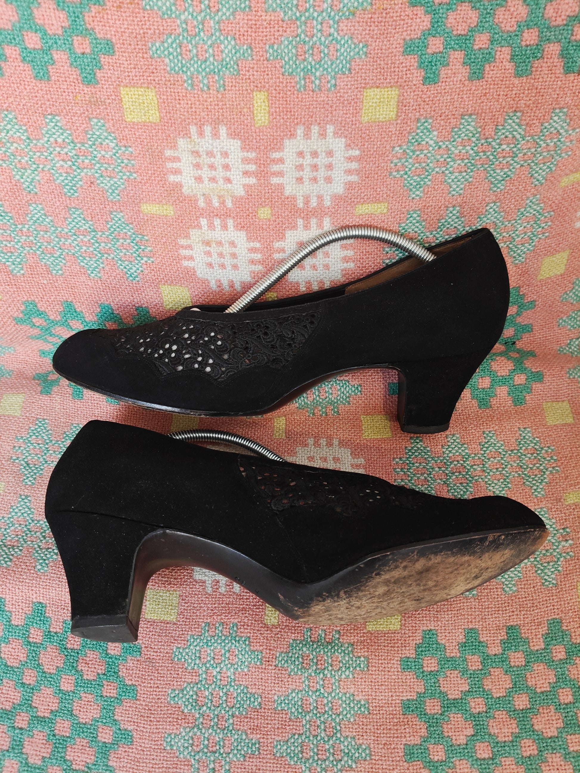A pair of black suede high-heeled vintage pumps with  floral patterns on the toe, displayed on a patterned pink, green, and yellow textile background.