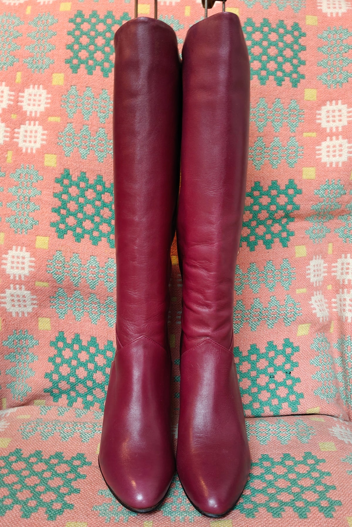 A pair of knee-high burgundy leather boots with pointed toes, displayed against a patterned pink, green, and white fabric background.