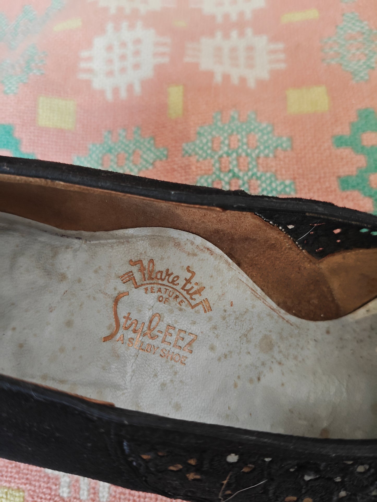 Close-up of the inside of a black leather shoe showing a worn beige insole with embossed text reading 'Mare Feature of StyleEZ AS A WAY SHOE', placed on a patterned pink and green textile surface.