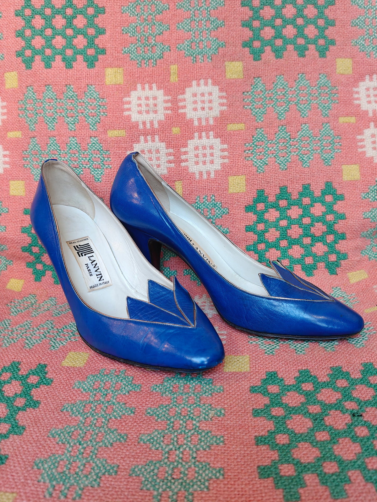 A pair of blue leather high-heeled pumps with pointed toes and geometric cutout details, displayed on a pink and green patterned textile background. The insole features a visible 'LANVIN PARIS' brand label.