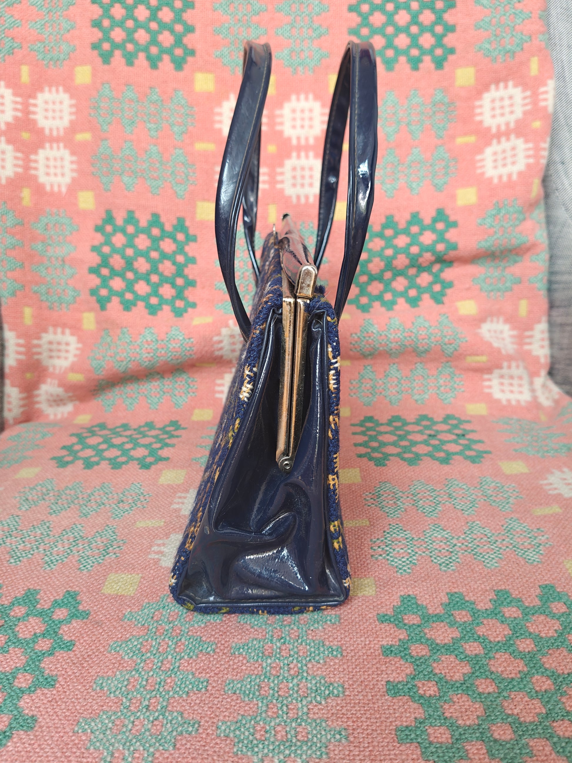 A navy blue patent leather handbag with  a gold-tone metal clasp, standing upright on a pink and green patterned textile surface.