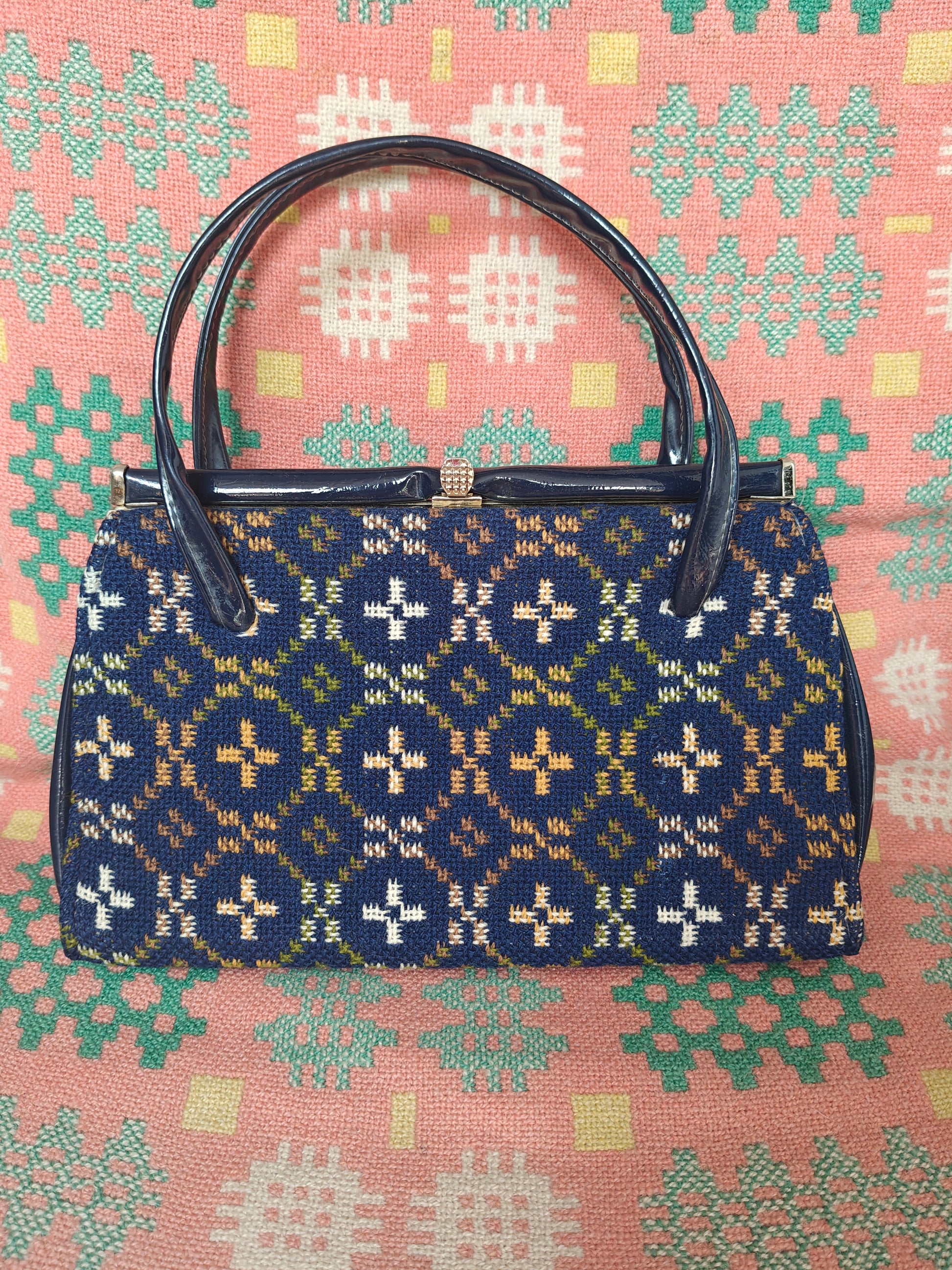 A vintage structured handbag with a navy blue woven pattern featuring cream, yellow, and green motifs, with dark brown leather handles and a gold-tone clasp, set against a pink patterned fabric background.