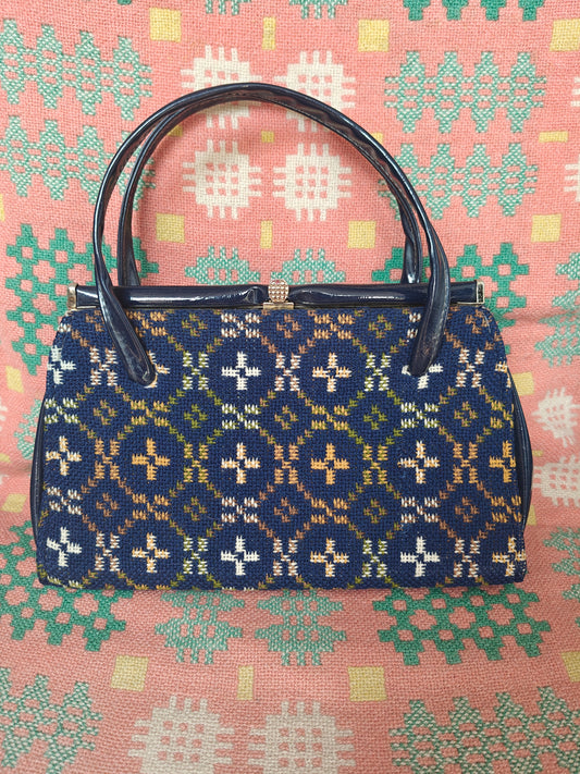 A vintage structured handbag with a navy blue woven pattern featuring cream, yellow, and green motifs, with dark brown leather handles and a gold-tone clasp, set against a pink patterned fabric background.