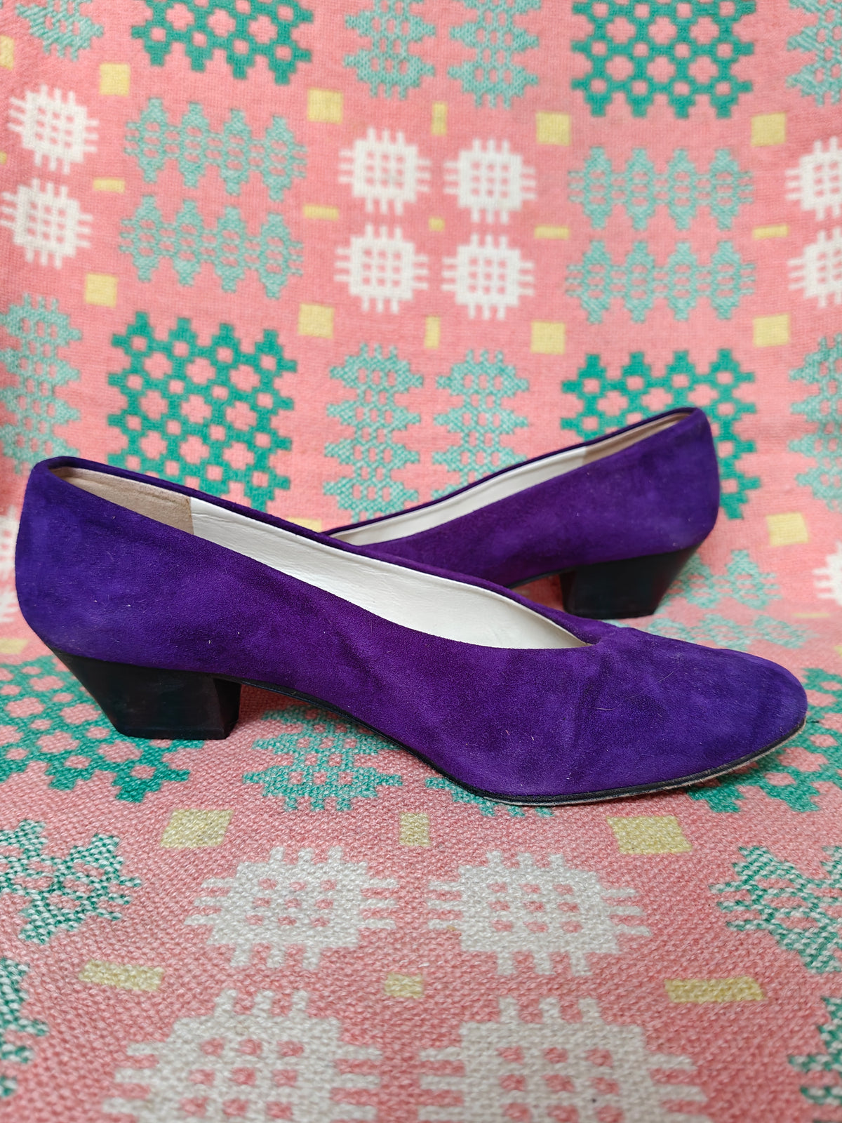 A pair of purple suede low-heel pumps with black block heels, displayed on a pink and green patterned woven rug.