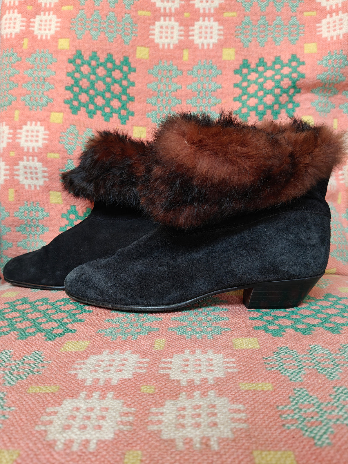Black suede ankle boots with a brown and black faux fur cuff, displayed on a pink and green patterned woven textile background.