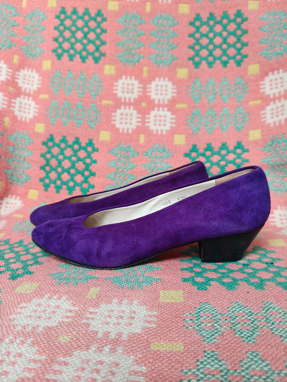 A pair of purple suede low-heel pumps with black block heels, displayed on a pink and teal patterned woven blanket.