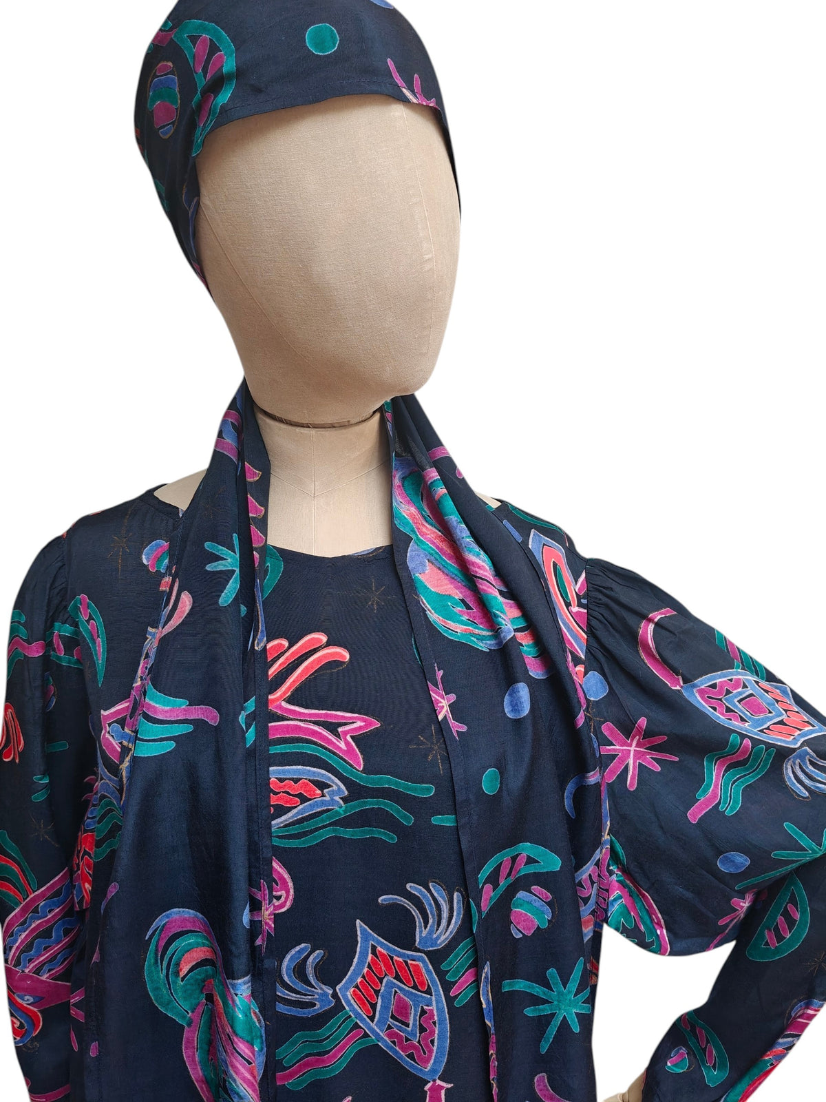 A mannequin wearing a navy blue outfit with a matching headscarf featuring a colourful paisley and planet pattern in pink, teal, and green.