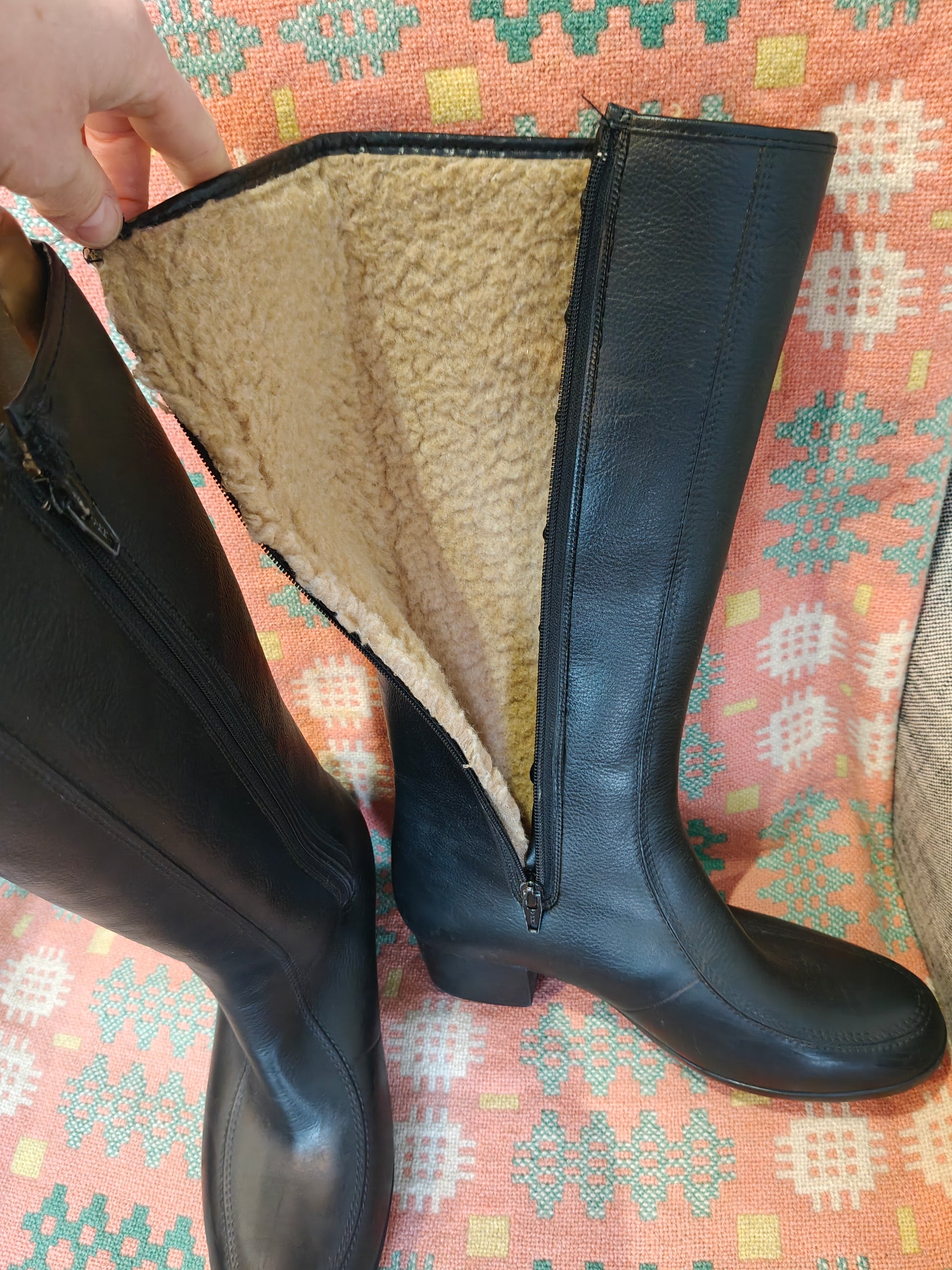 Black knee-high boot with fur lining held by a hand on a patterned fabric background
