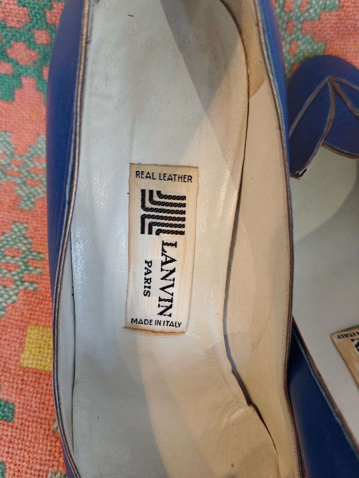 A close-up of the interior of a blue Lanvin shoe, showing the brand label 'LANVIN PARIS' on the cream-colored footbed, resting on a colorful patterned textile background.