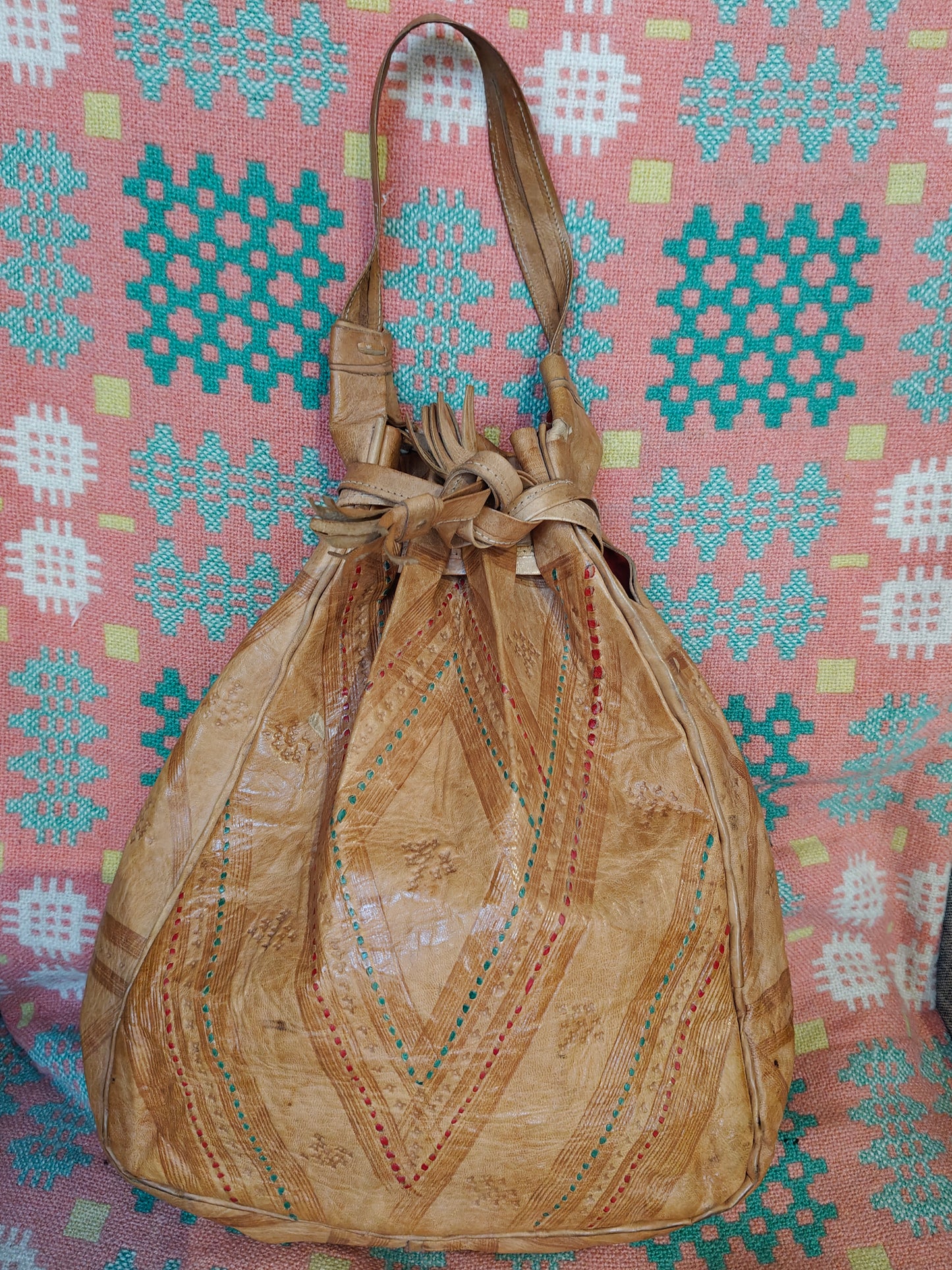 A tan leather drawstring bucket bag with embroidered geometric patterns, displayed against a pink and teal patterned textile background.