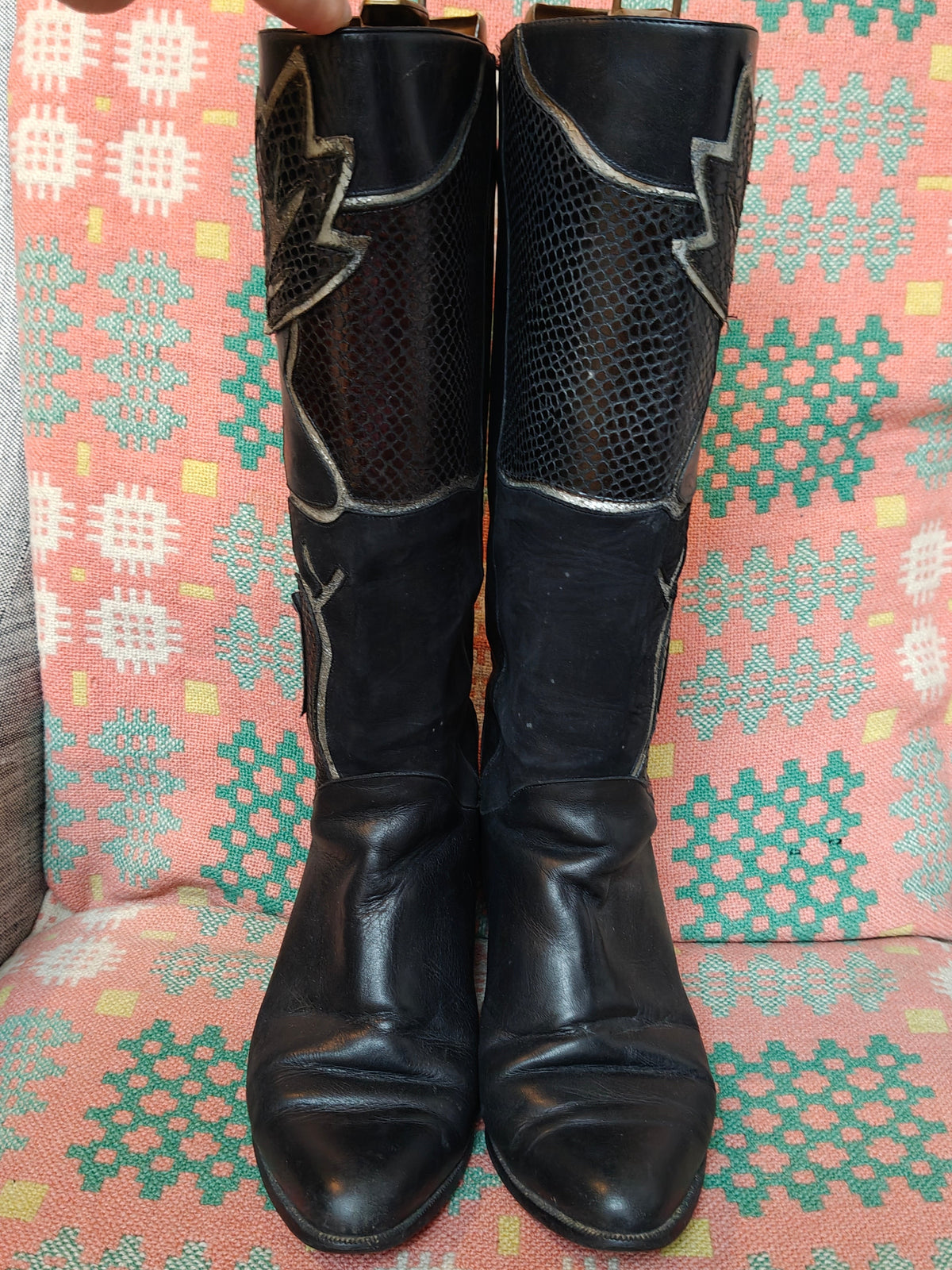 A pair of black leather knee-high western boots with textured mesh panels and silver decorative stitching, shown standing on a patterned pink, green, and white chair with a hand pointing at them.