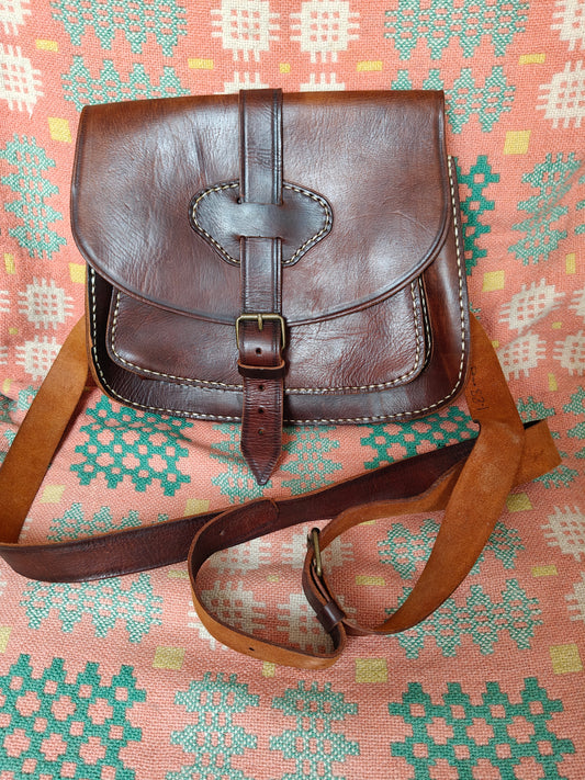 A brown leather crossbody bag with a front flap, buckle strap, and long shoulder strap, laid flat on a patterned pink and green textile background.