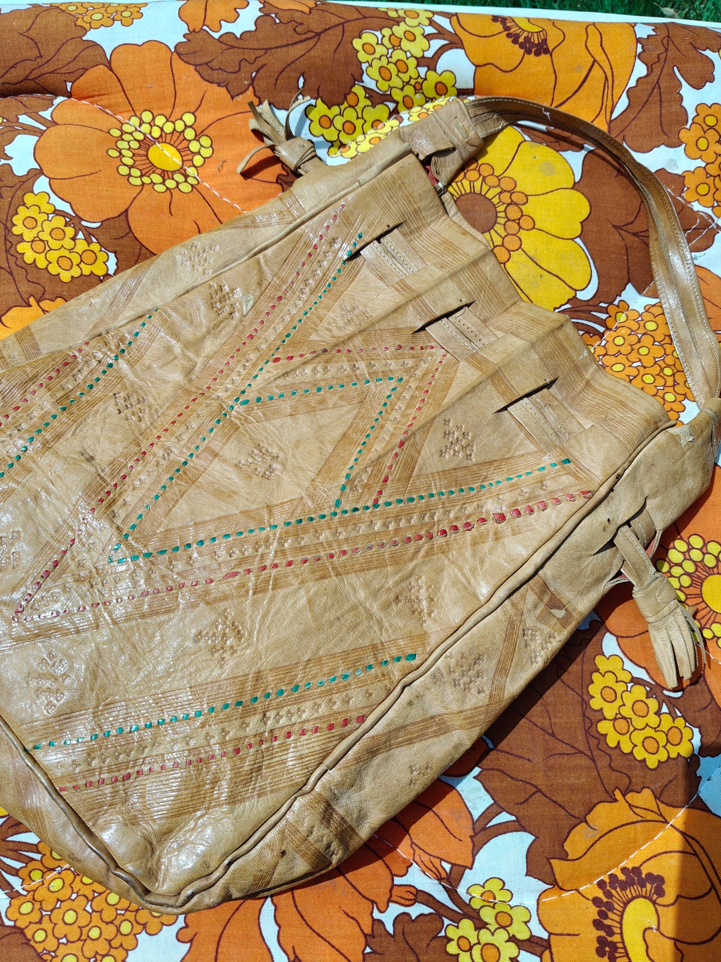 A vintage tan leather shoulder bag with geometric patterns in red, green, and blue, laid flat on a retro floral orange and yellow blanket.