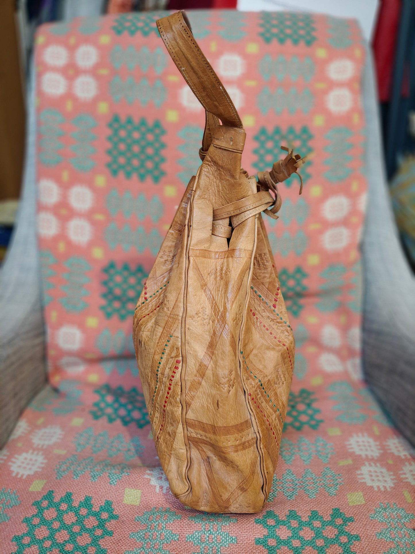 A tan leather bag with geometric stitched patterns, shown from the side on a patterned pink and teal chair.