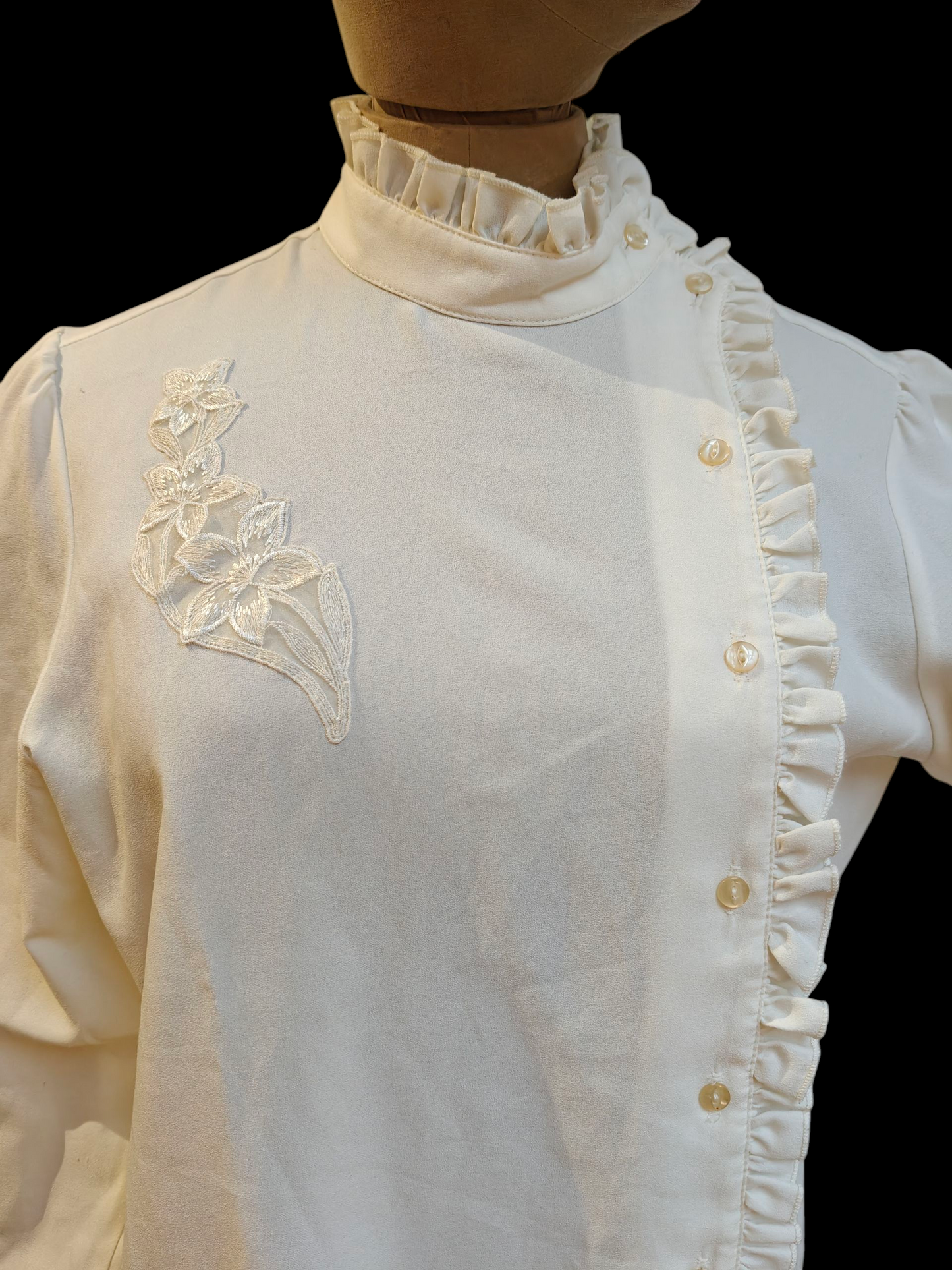 White blouse with floral embroidery and ruffled details on a mannequin against a black background