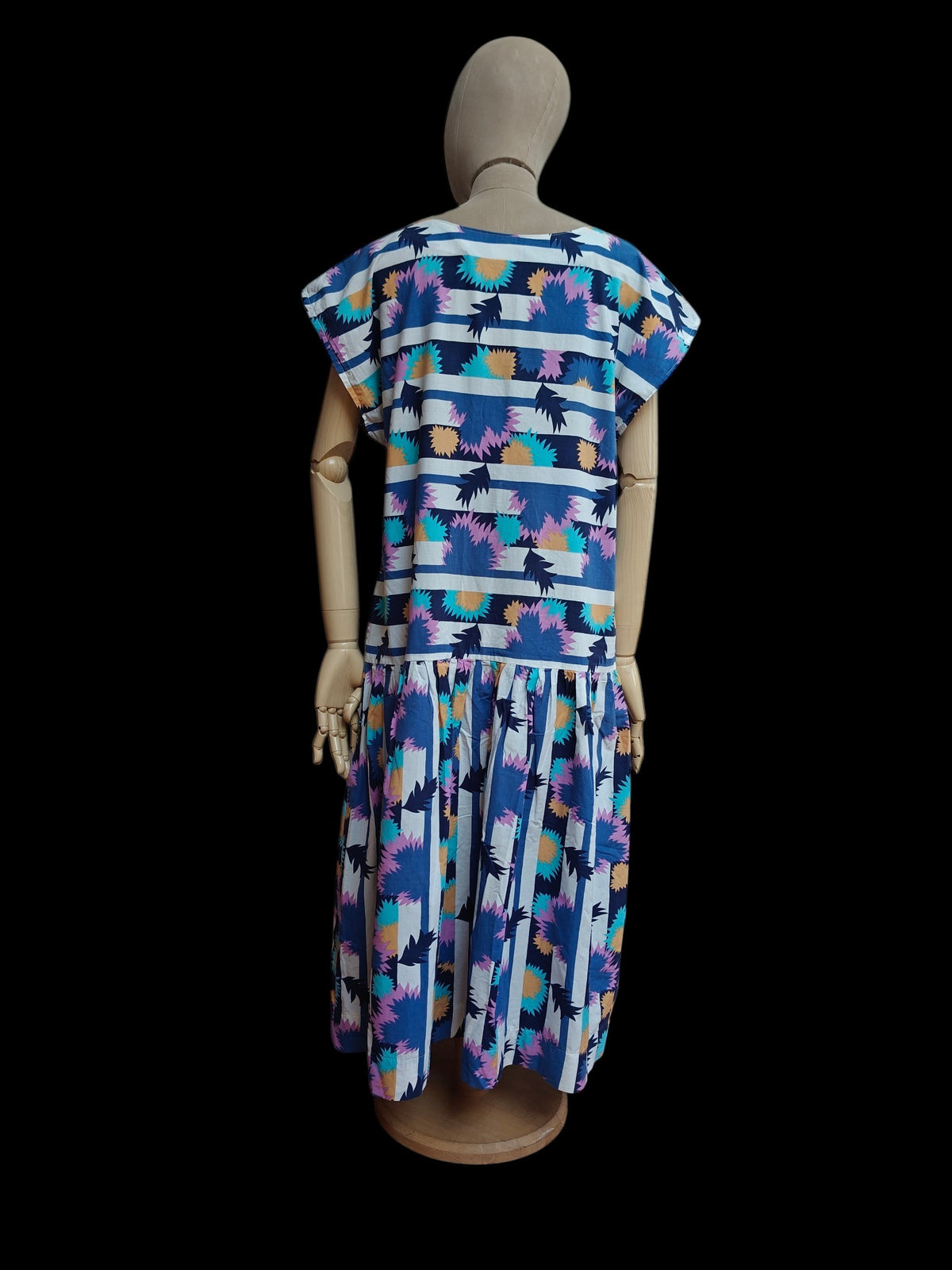 A short-sleeve 1980's dress displayed on a wooden mannequin from the back, featuring a blue and white striped top with colorful palm tree prints and a matching pleated skirt, set against a solid black background.