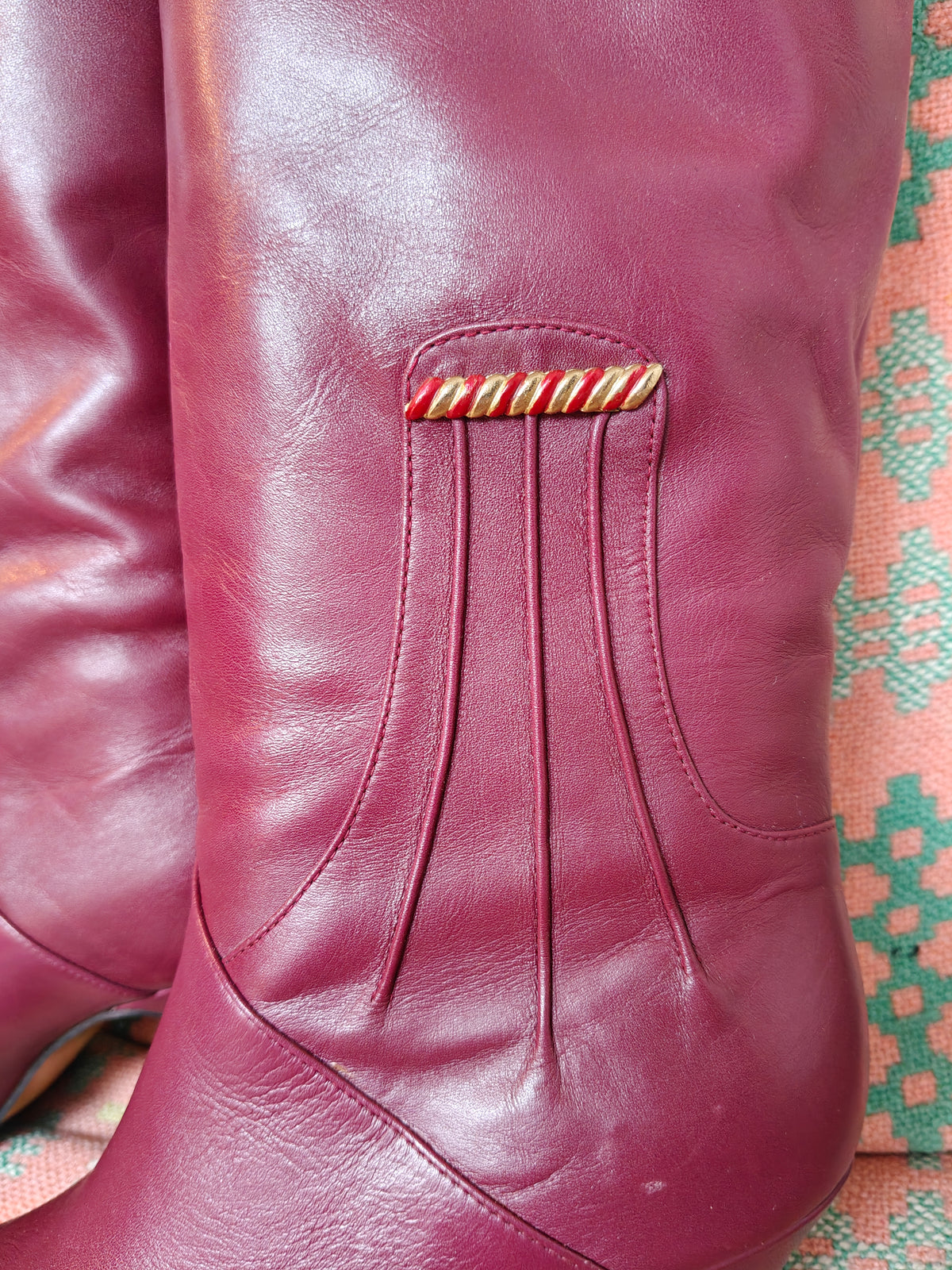 A close-up of a burgundy leather boot featuring vertical pleats and a decorative red and yellow striped leather accent piece near the top.